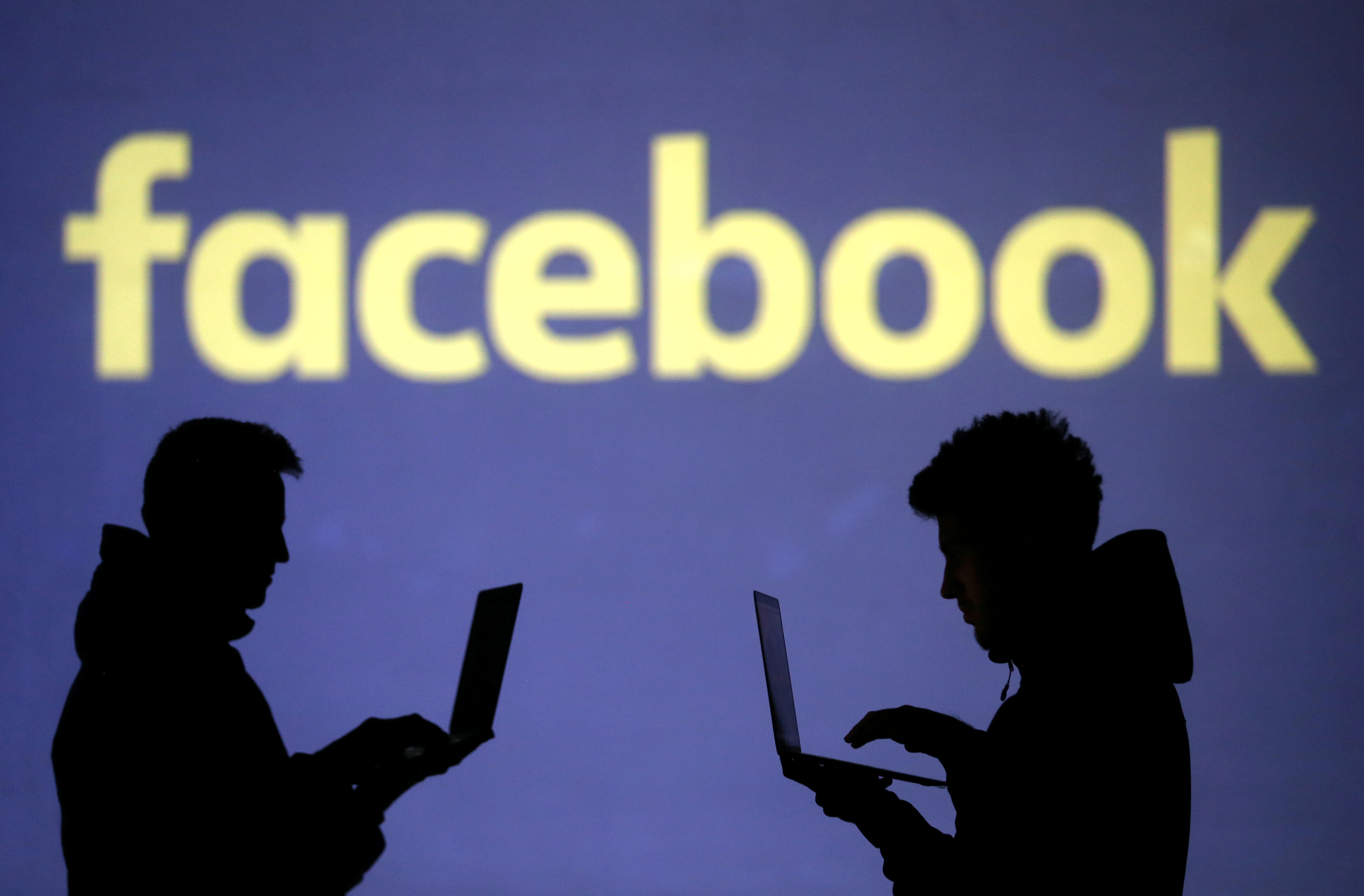 Silhouettes of laptop users are seen next to a screen projection of Facebook logo in this picture illustration