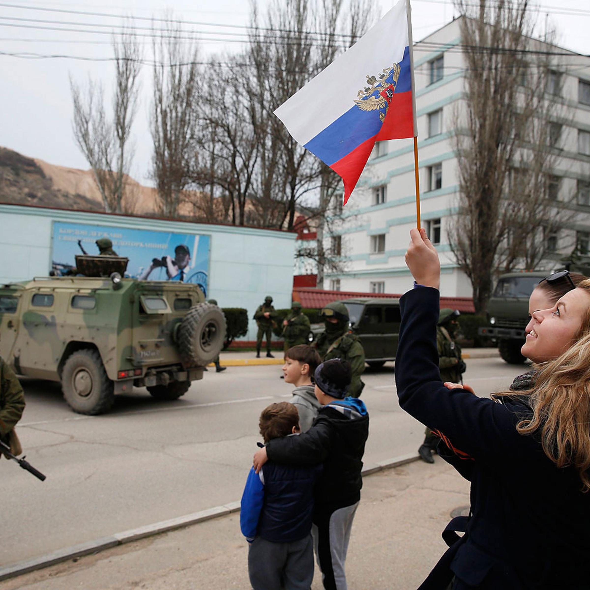 A woman waves a Russian flag as armed servicemen wait near Russian army vehicles outside a Ukrainian border guard post in the Crimean town of Balaclava