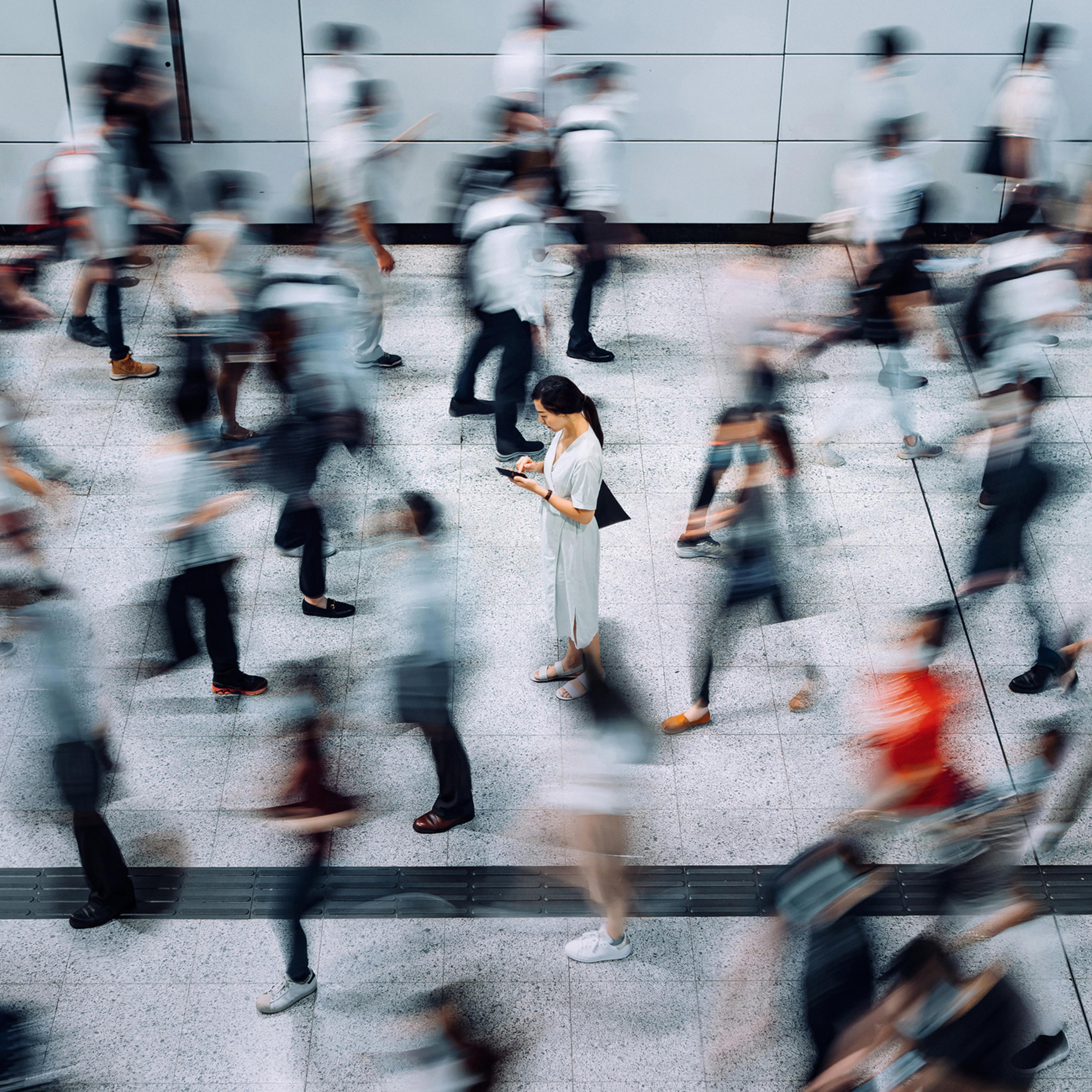 Young Asian woman using smartphone surrounded by commuters rushing by in subway station during office peak hours in the city