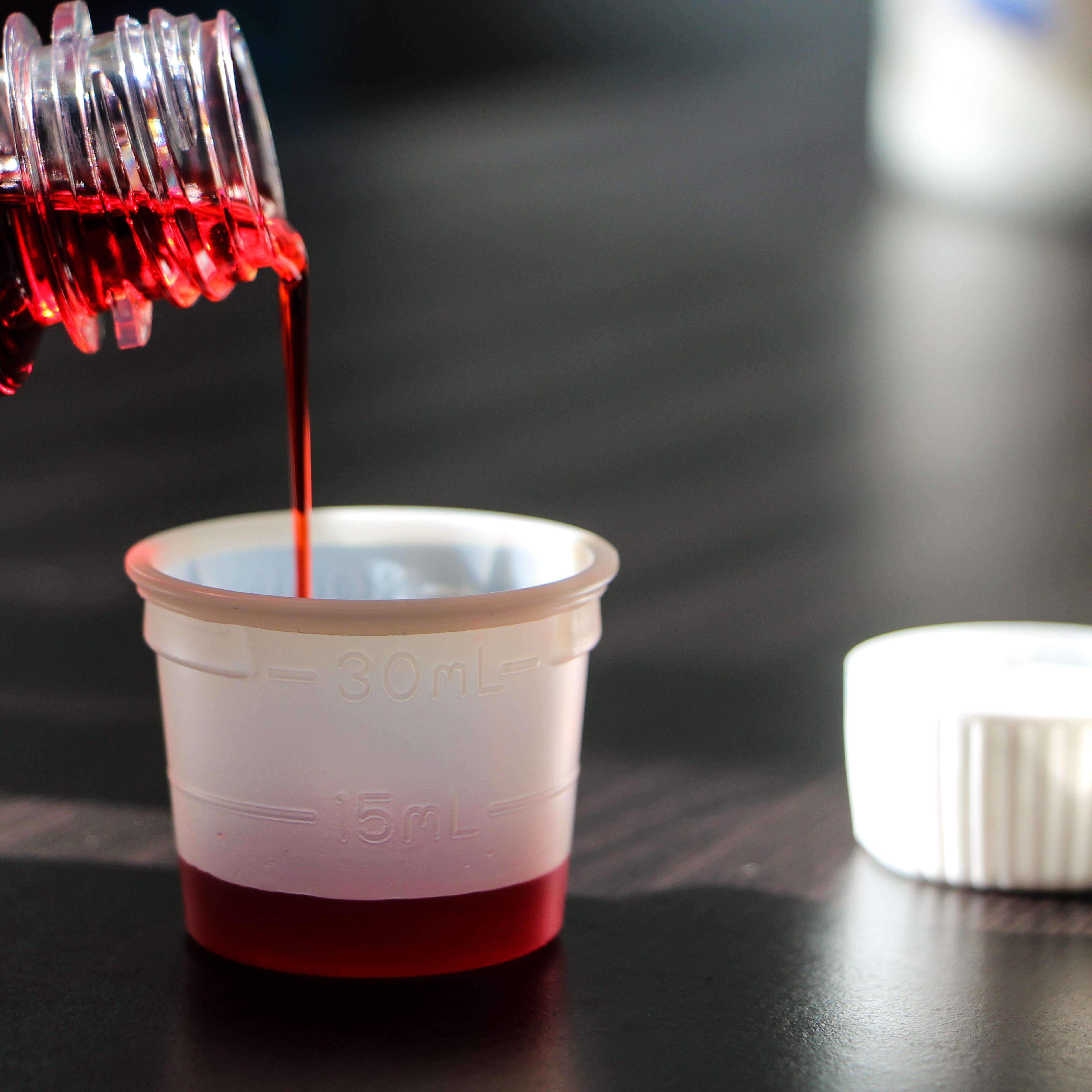 Close-Up Of Bottle Pouring Cough Syrup In Container On Table
