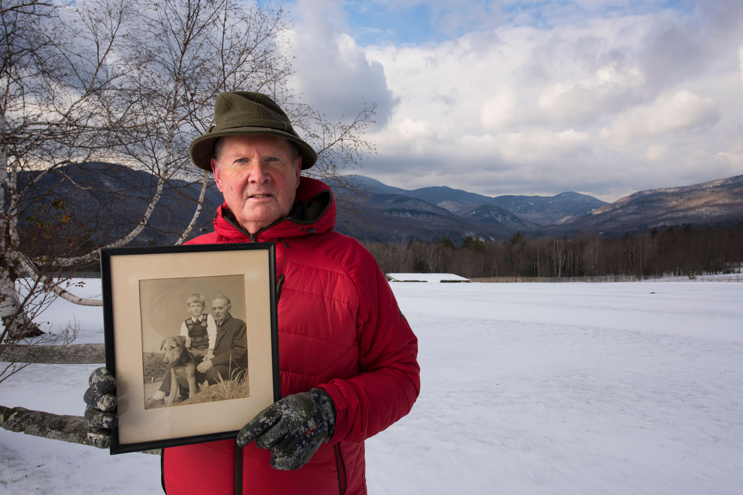Portrait of Johannes von Trapp at the Trapp Family Lodge, alongside a field where music is performed in warmer weather. He is holding a portrait of his father and himself.
