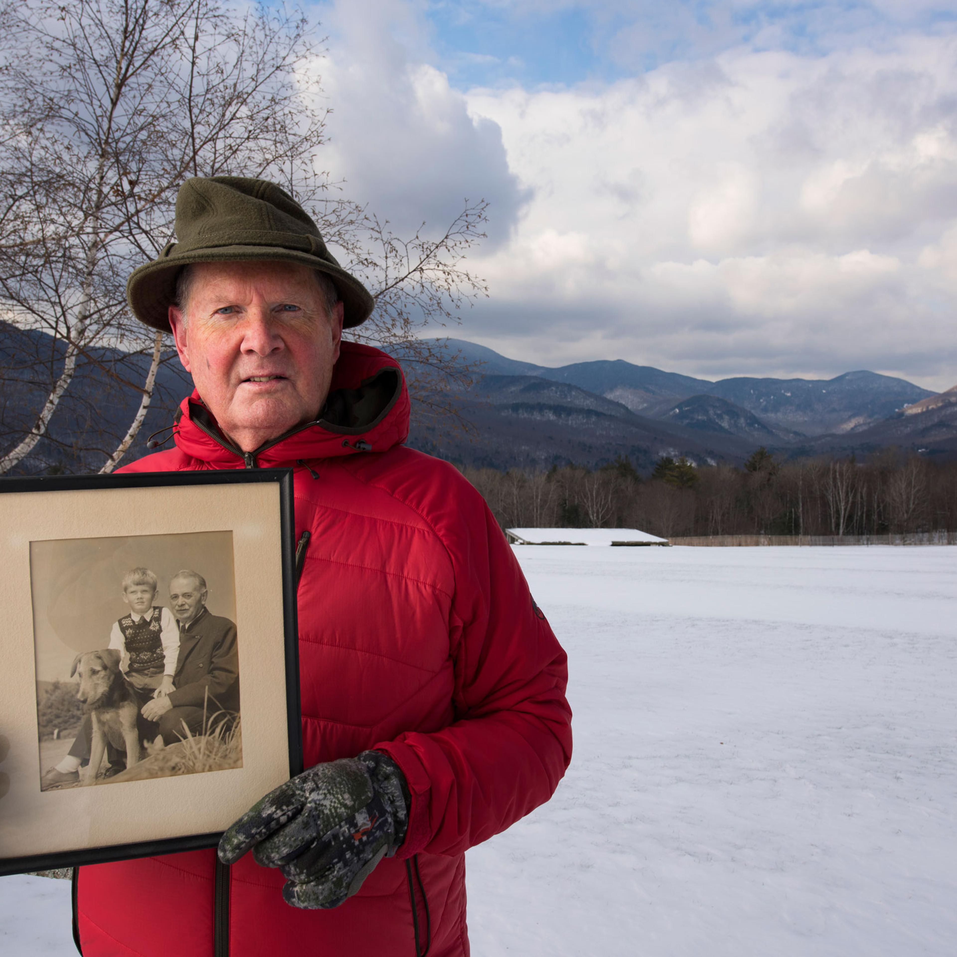 Portrait of Johannes von Trapp at the Trapp Family Lodge, alongside a field where music is performed in warmer weather. He is holding a portrait of his father and himself.