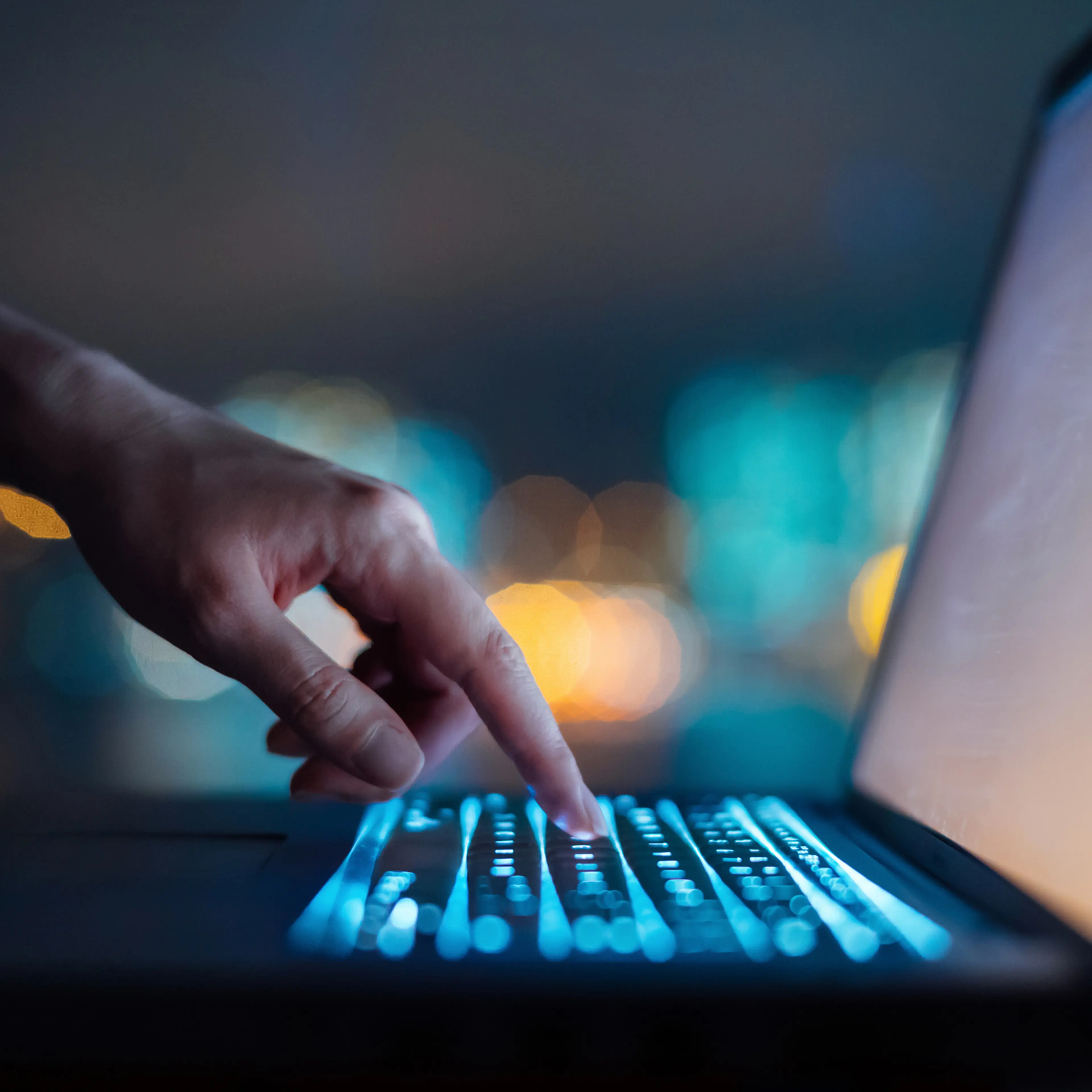 Close up of woman's hand typing on computer keyboard in the dark against colourful bokeh in background, working late on laptop at home