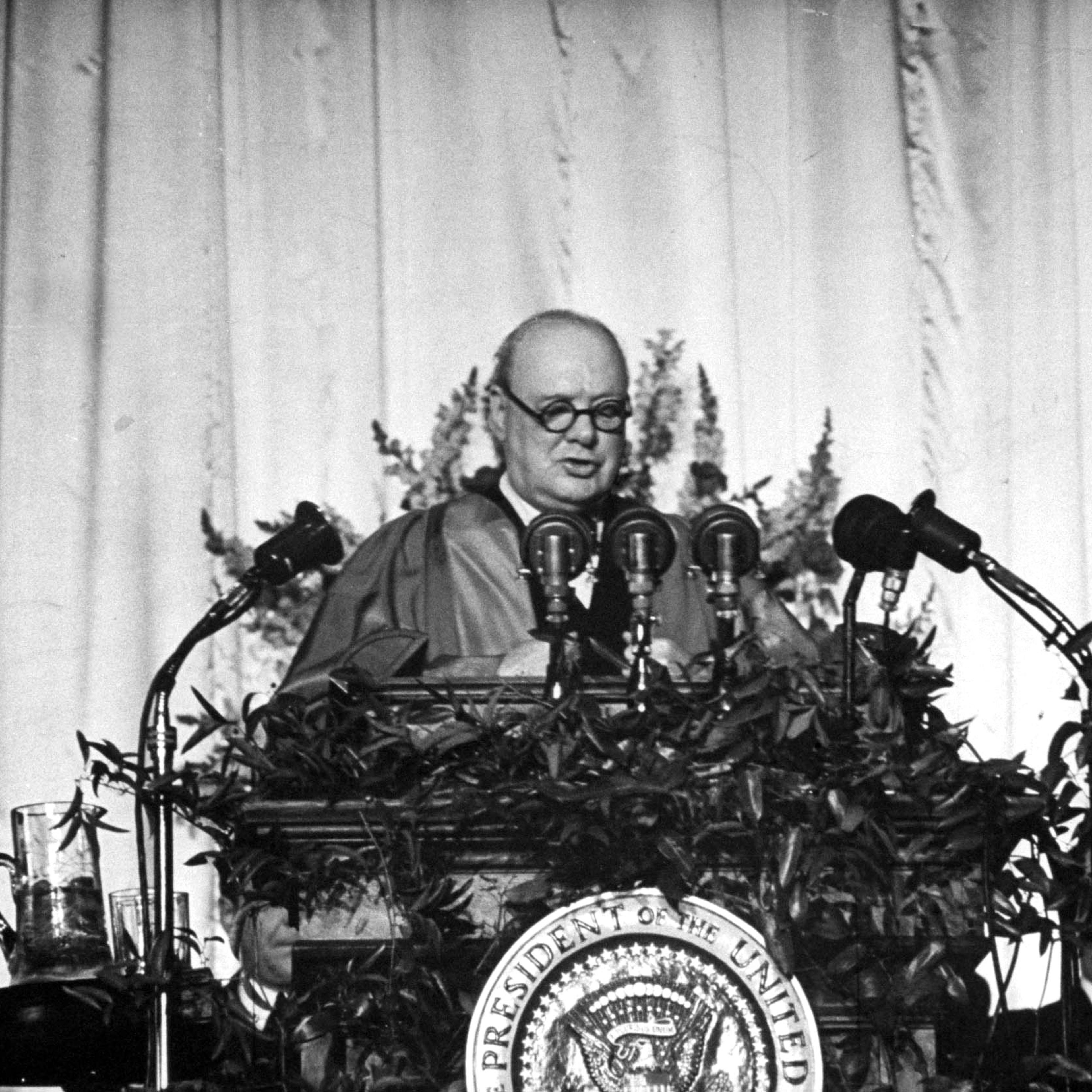 Churchill speaks in the gym from a rostrum decorated with shrubs and radio microphones. His small audience sat on hard, borrowed bleachers.