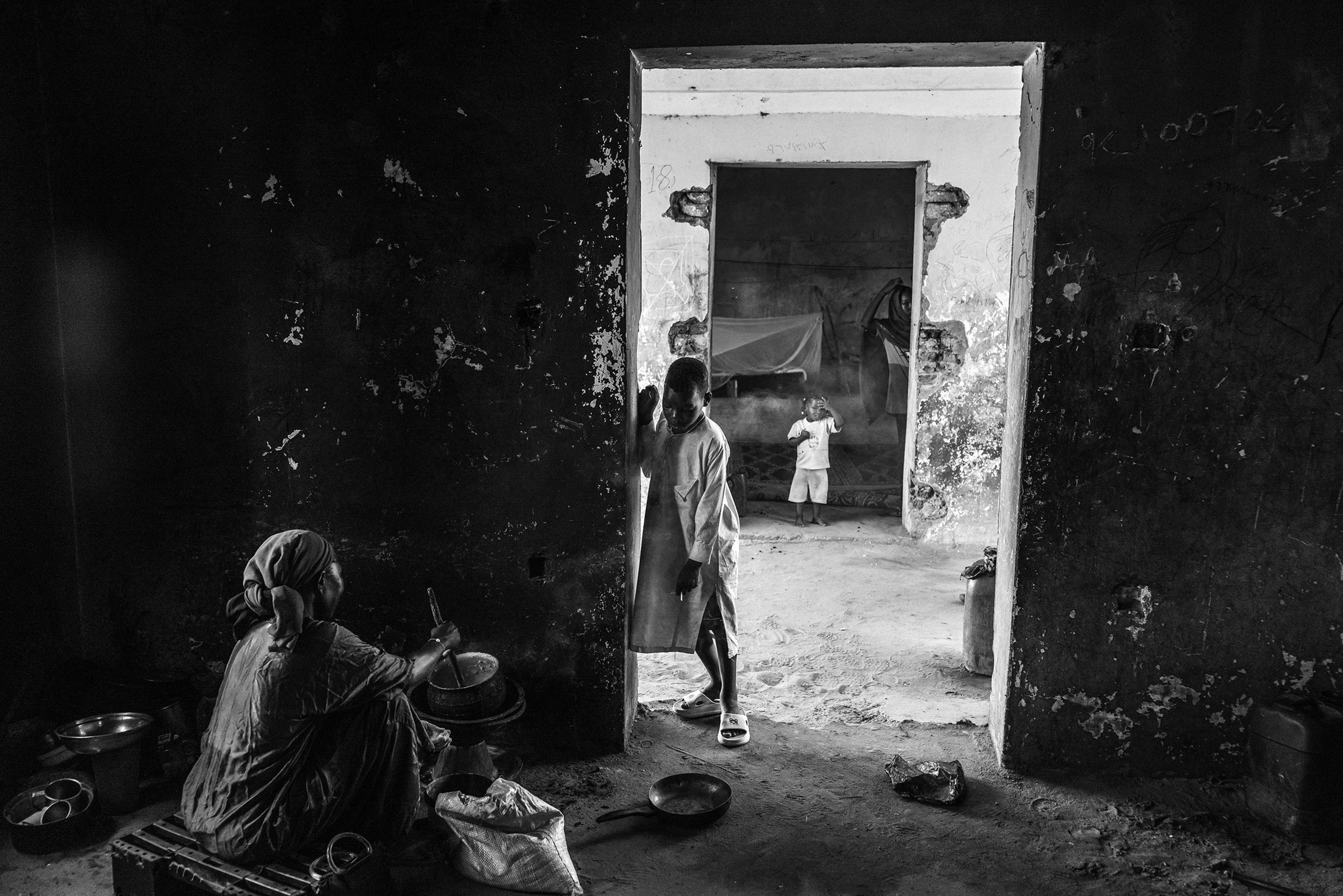 A woman cooks in the shadows of a former orphanage while children move through adjoining rooms, now converted into living quarters for families displaced from Khartoum. The abandoned compound on the outskirts of Al Junaynah shelters dozens who fled Sudan