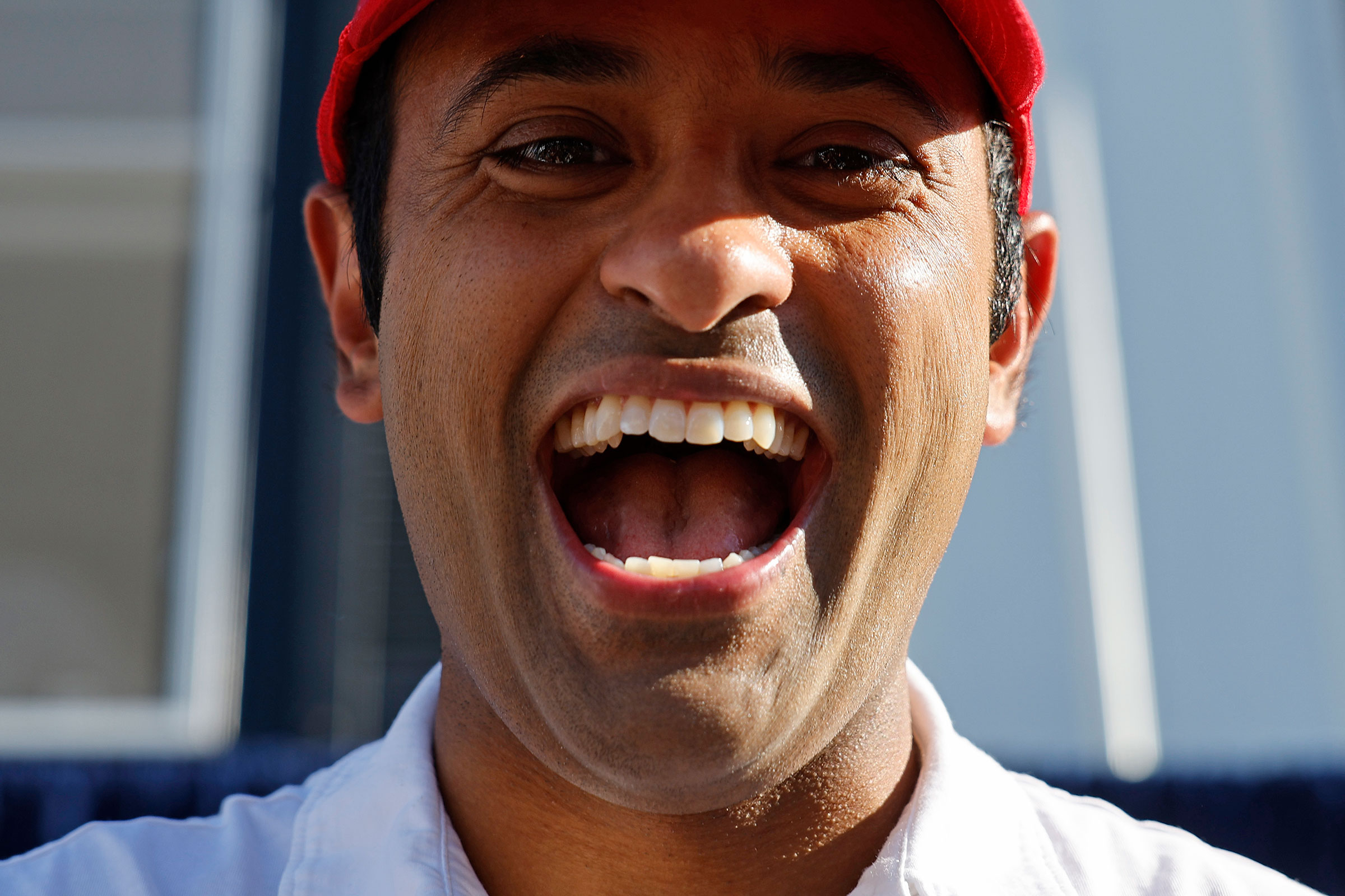 A close up portrait of Vivek Ramaswamy with a wide smile in a red had and white polo