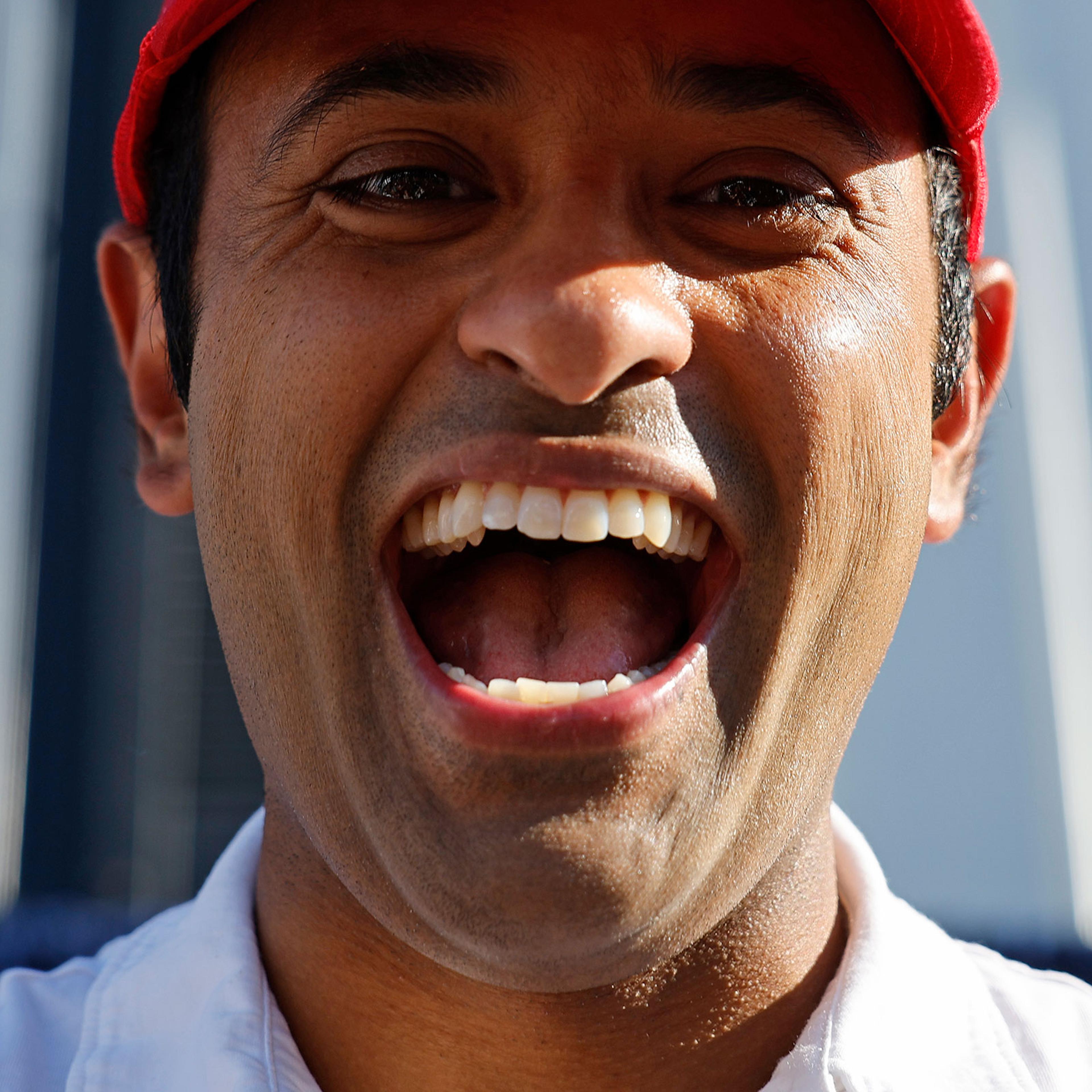 A close up portrait of Vivek Ramaswamy with a wide smile in a red had and white polo