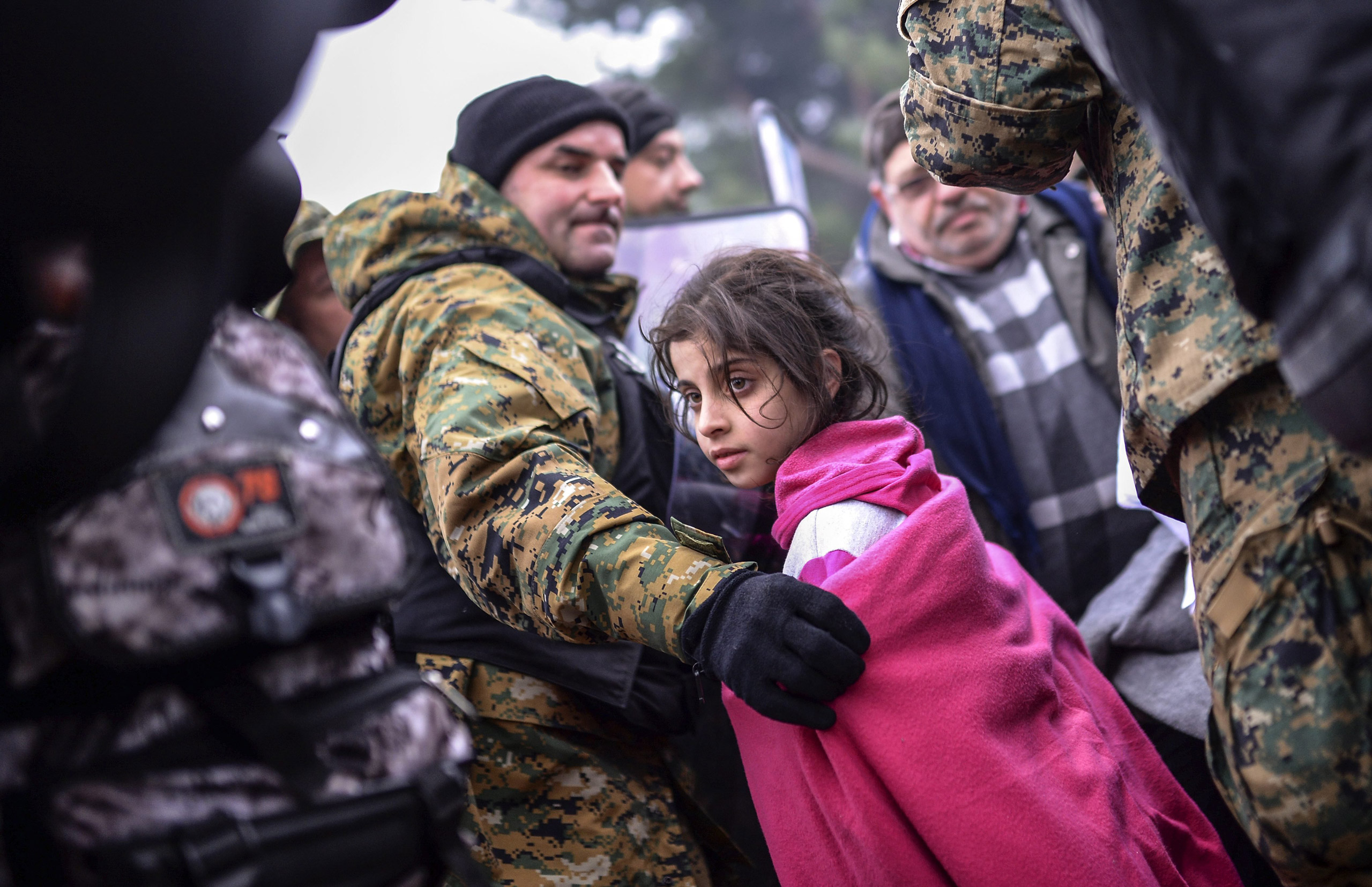 epa05034123 A Macedonian police officer helps a girl to pass between the cordon as migrants and refugees wait to cross the Greek-Macedonian border after Macedonia has started granting passage only to refugees from Syria, Iraq and Afghanistan, near Gevege