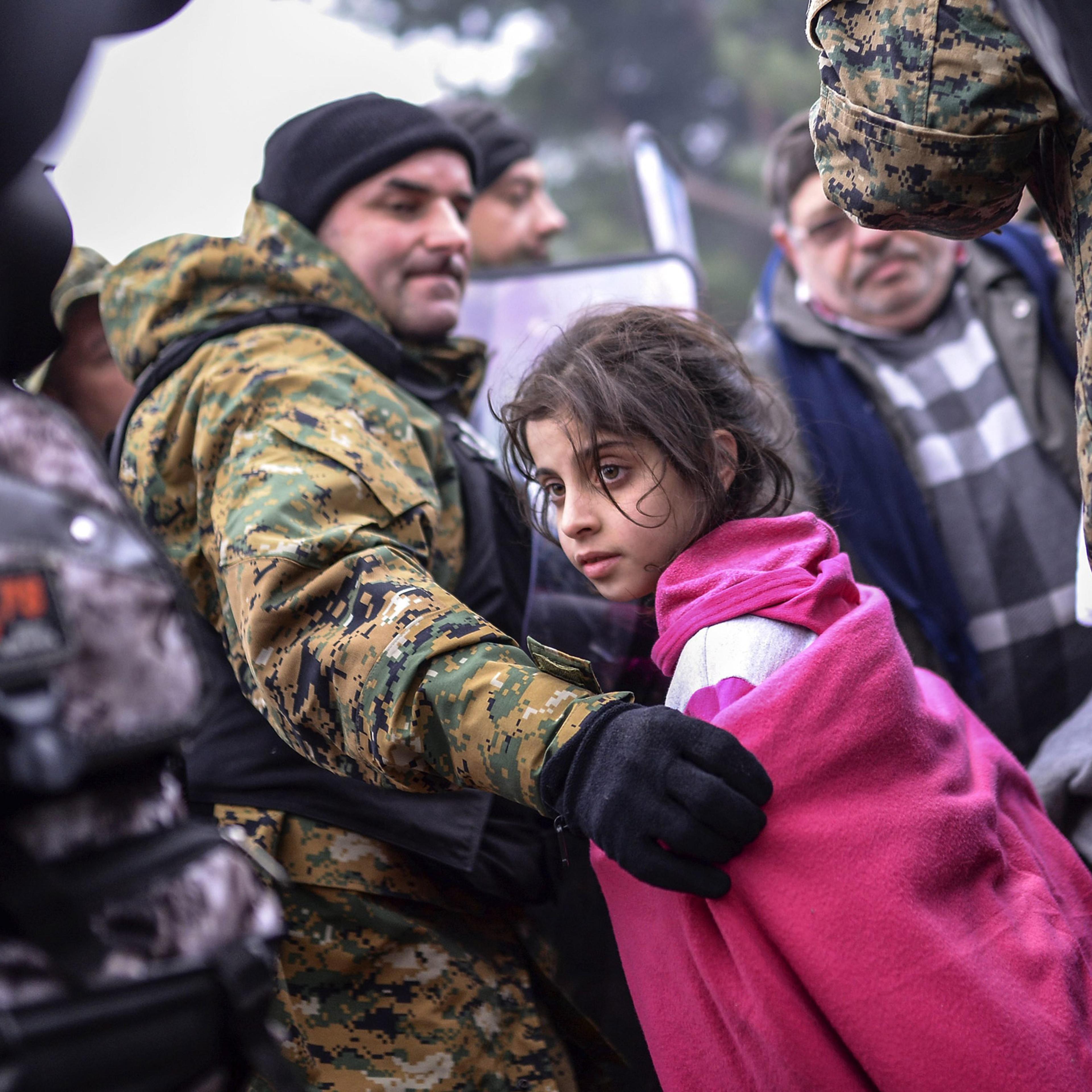 epa05034123 A Macedonian police officer helps a girl to pass between the cordon as migrants and refugees wait to cross the Greek-Macedonian border after Macedonia has started granting passage only to refugees from Syria, Iraq and Afghanistan, near Gevege