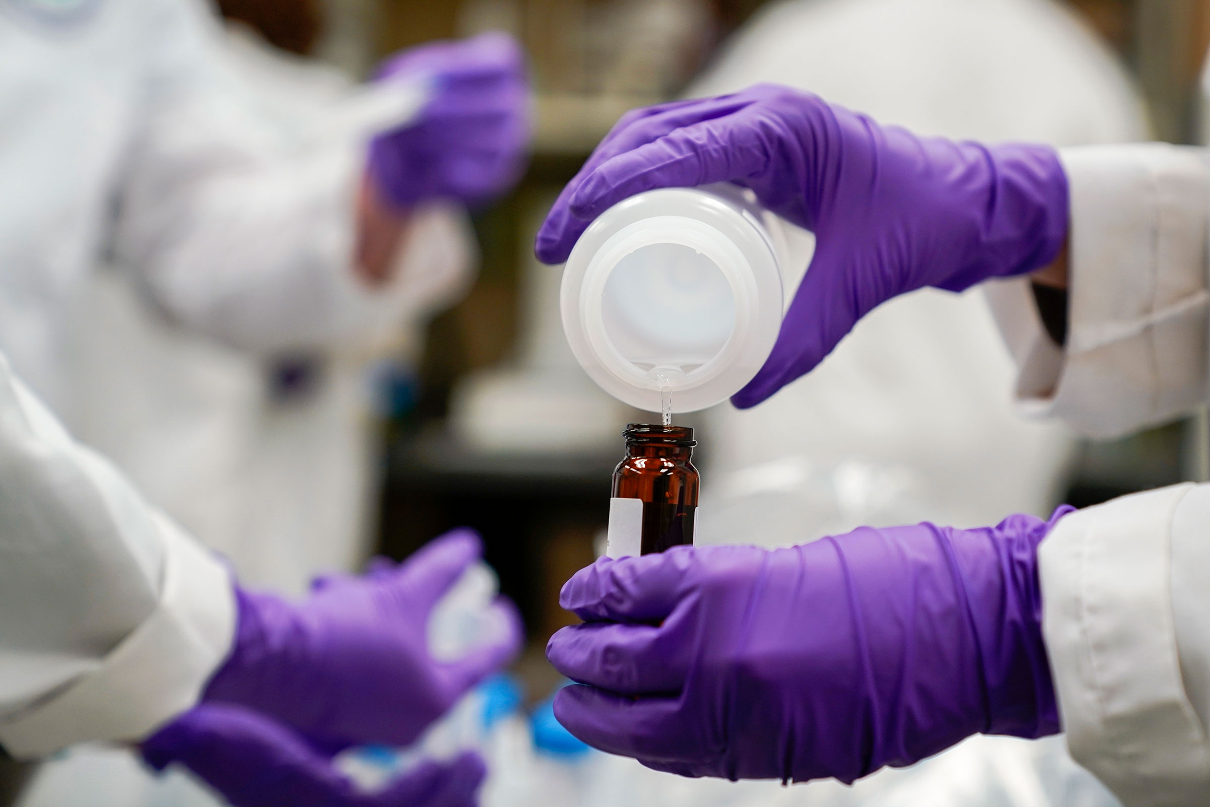 A researcher with rubber gloves pours a water sample into a smaller glass container for experimentation as part of drinking water and PFAS research