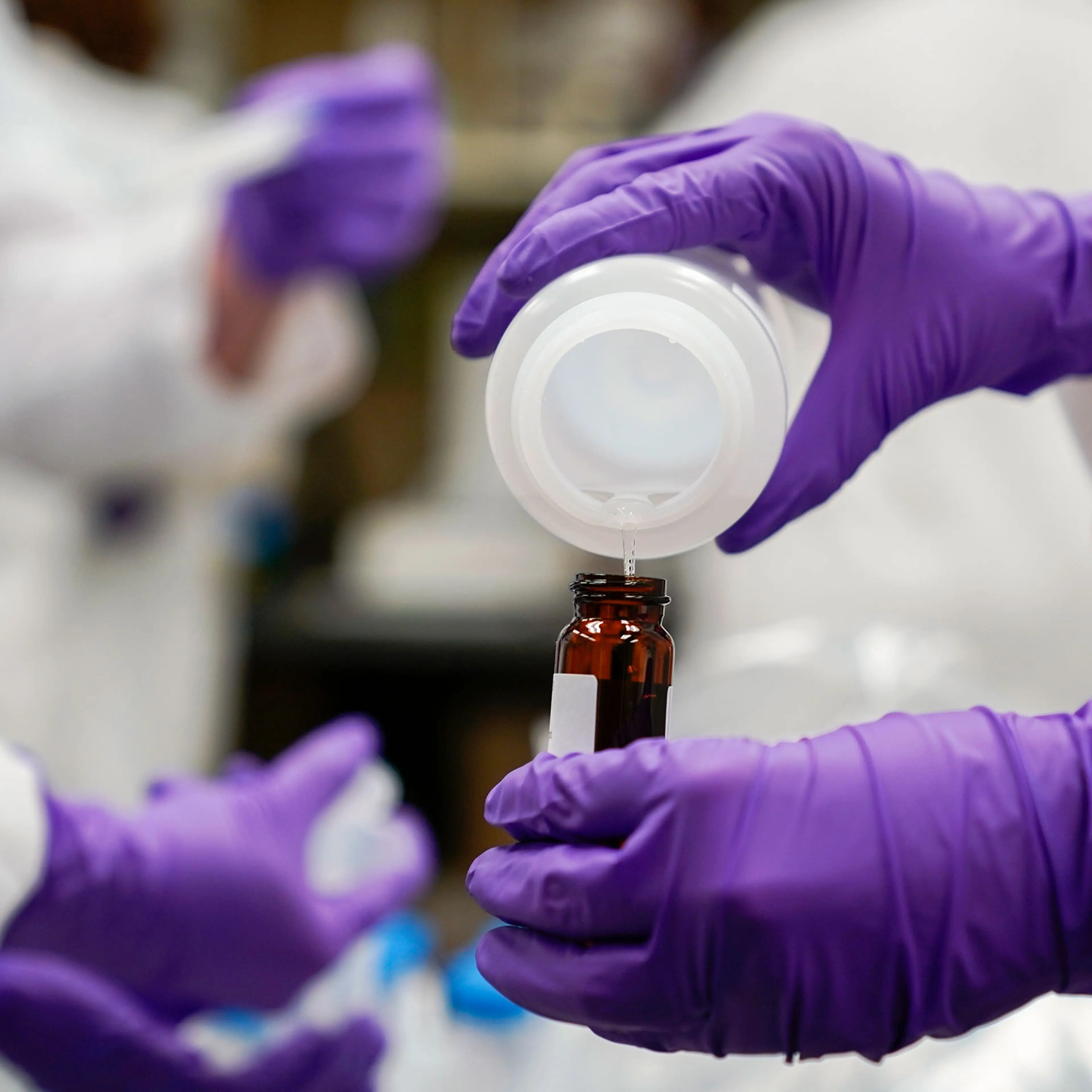 A researcher with rubber gloves pours a water sample into a smaller glass container for experimentation as part of drinking water and PFAS research
