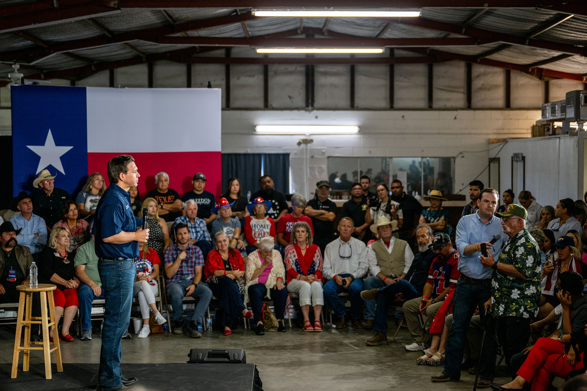DeSantis stands on a small stage facing a supporter among a crowd of people sitting around him who is asking a question into a microphone