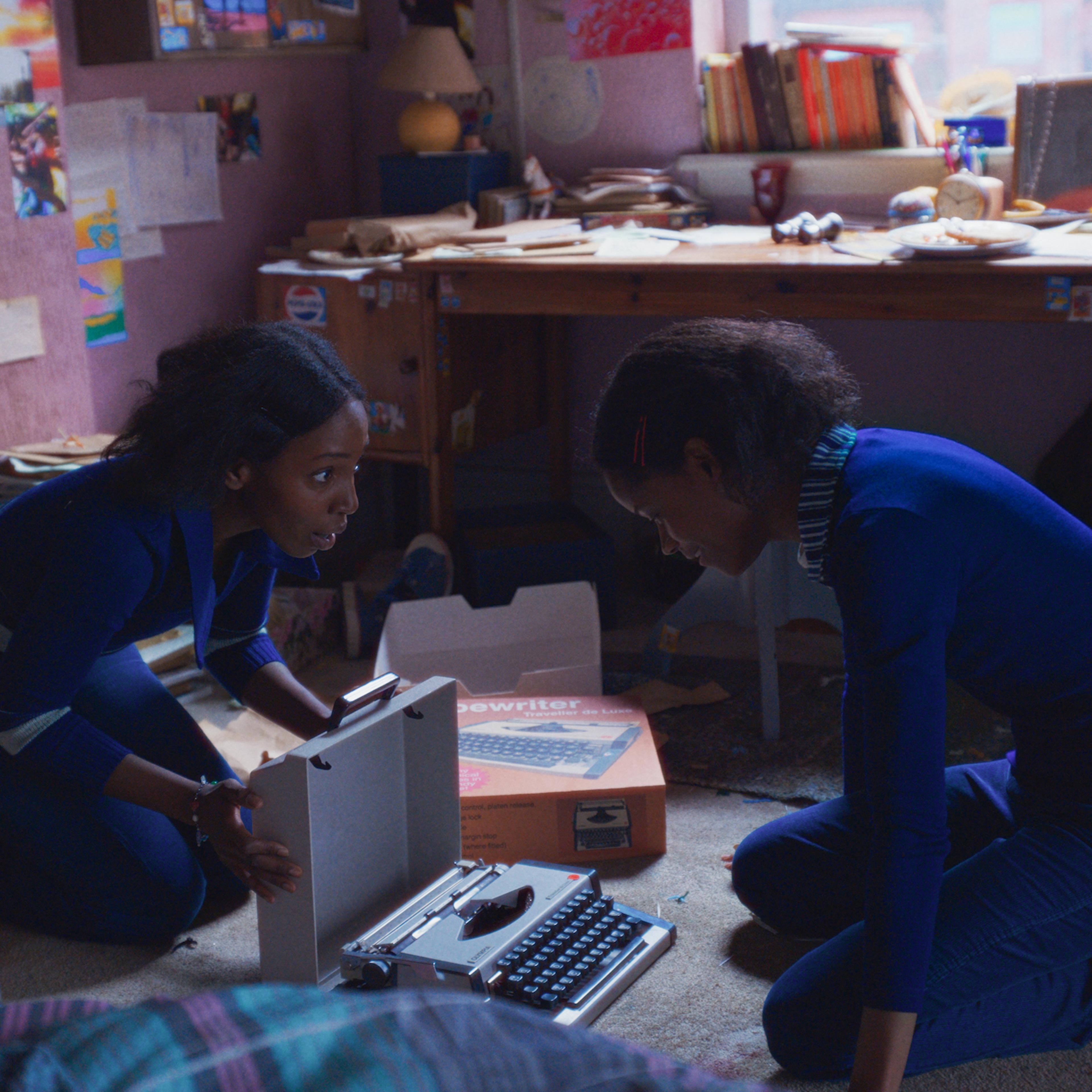 Tamara Lawrance as Jennifer Gibbons and Letitia Wright as June Gibbons open a new typewriter from its box in their bedroom.
