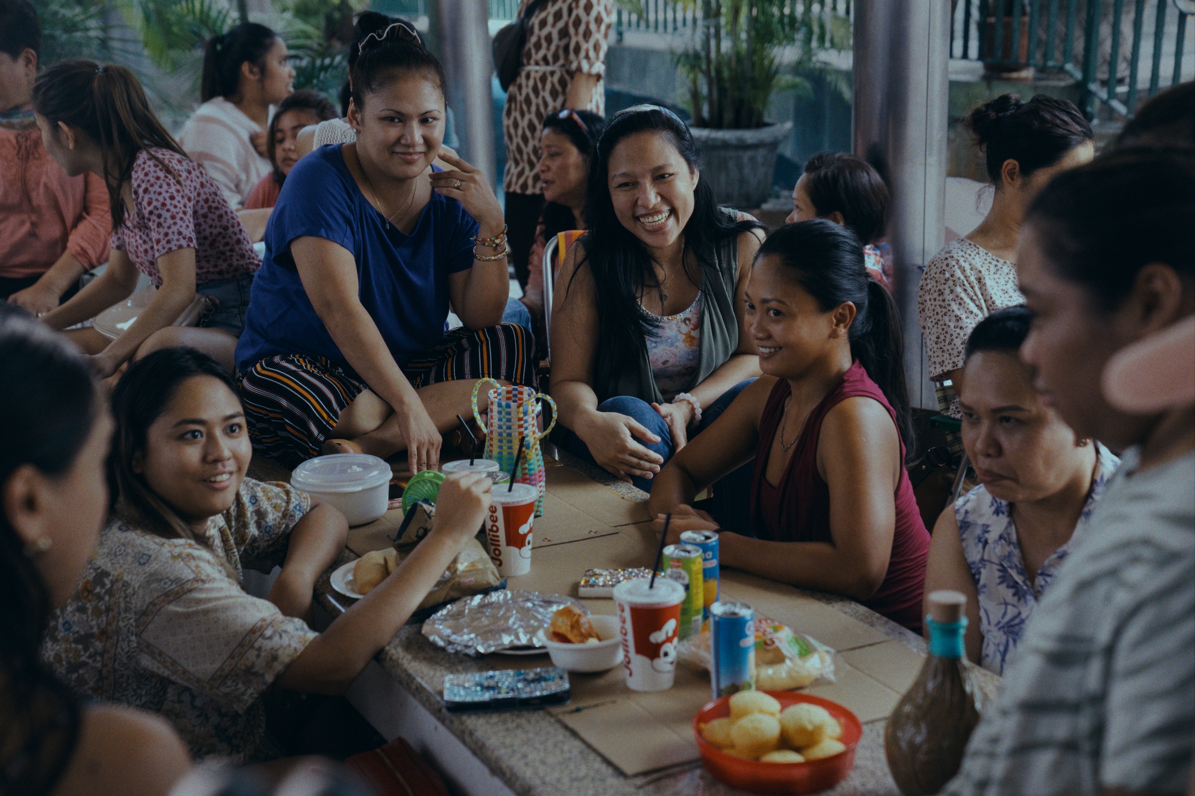 A circle of "helpers," including Puri (Amelyn Pardenilla), center, gather to hang out, eat, and gossip on their day off.