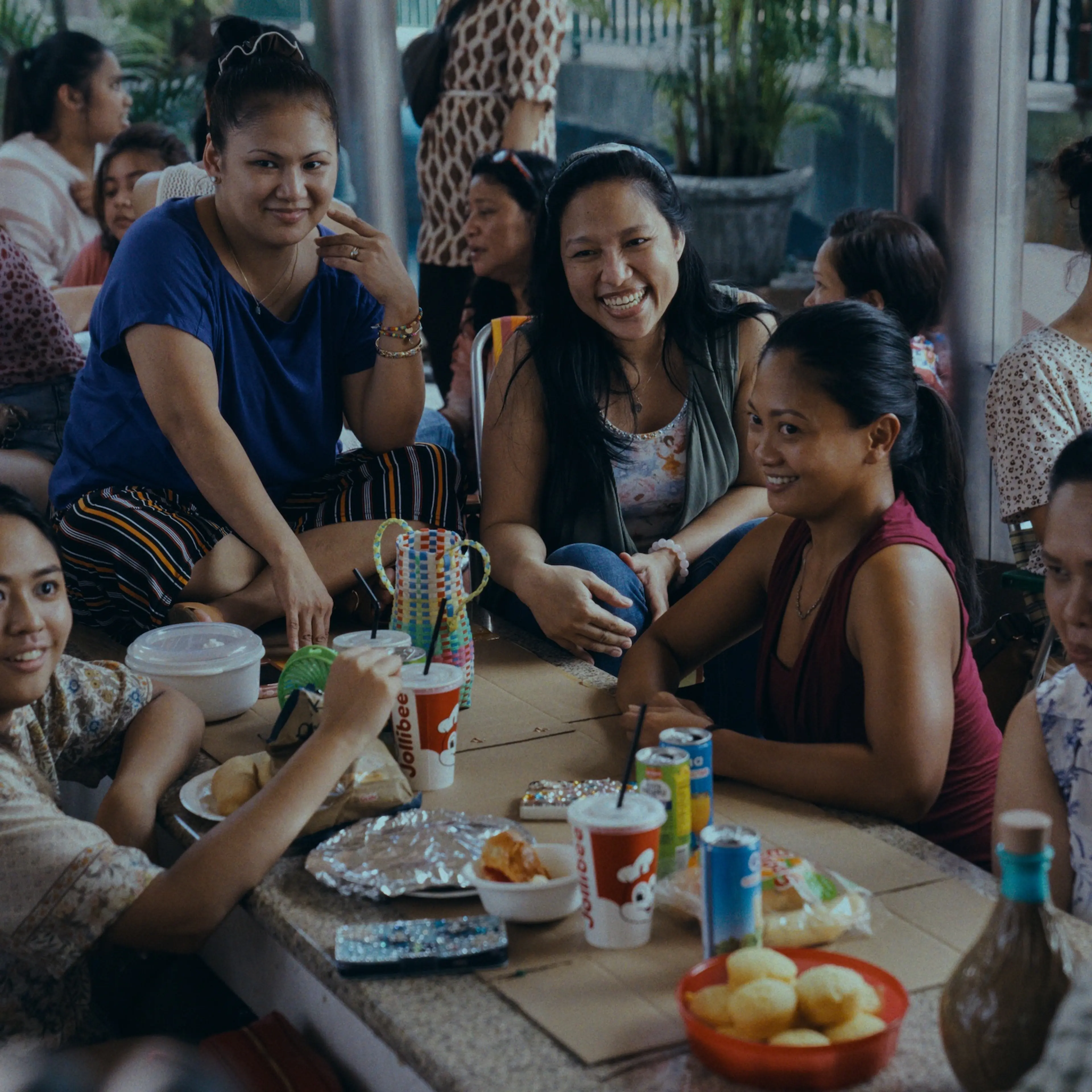 A circle of "helpers," including Puri (Amelyn Pardenilla), center, gather to hang out, eat, and gossip on their day off.