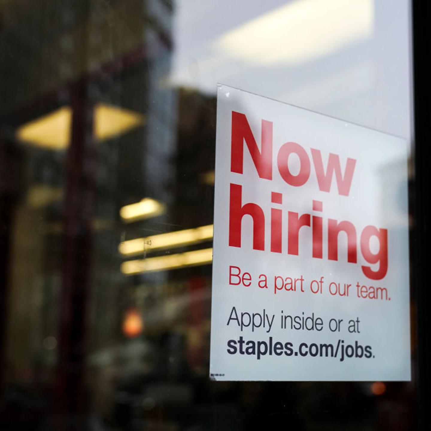 A 'now hiring' sign hangs on the door of a Staples store