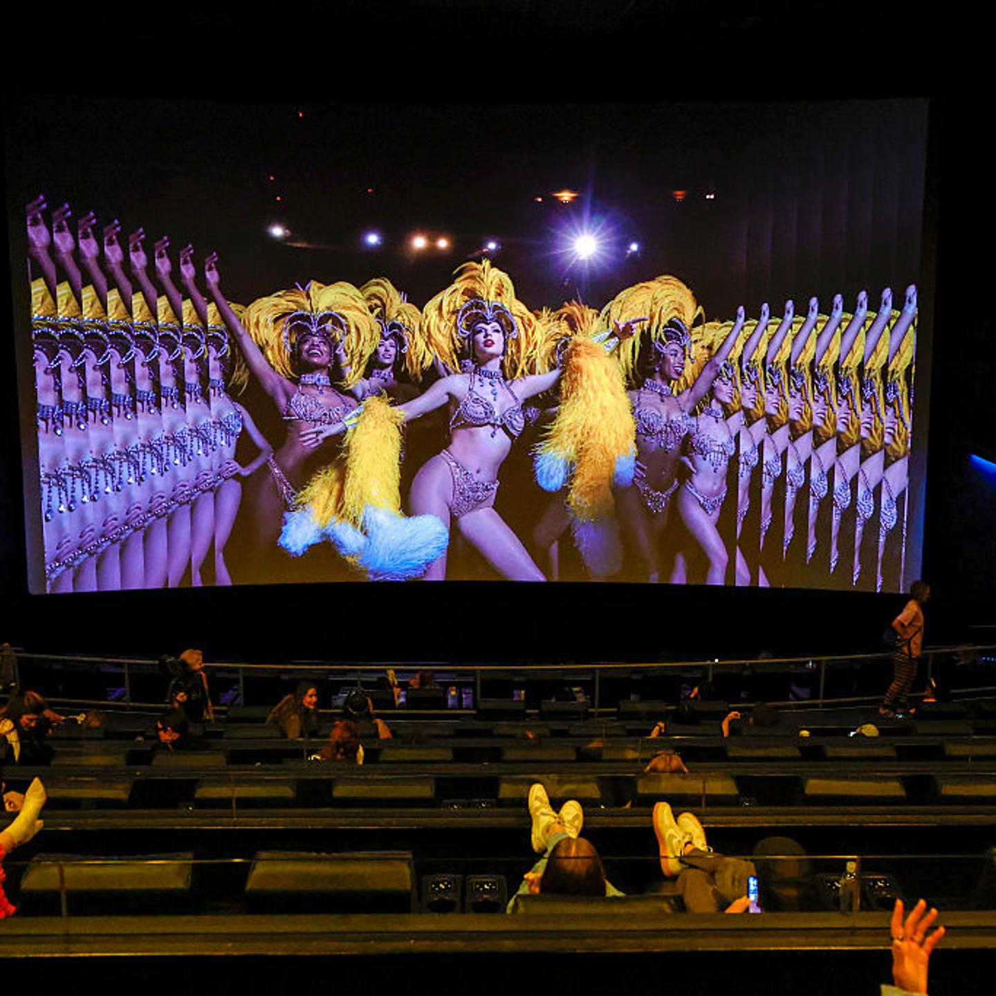 In a darkened movie theater, audience members kick up their feet and watch Taylor Swift dressed in yellow feathers on screen.