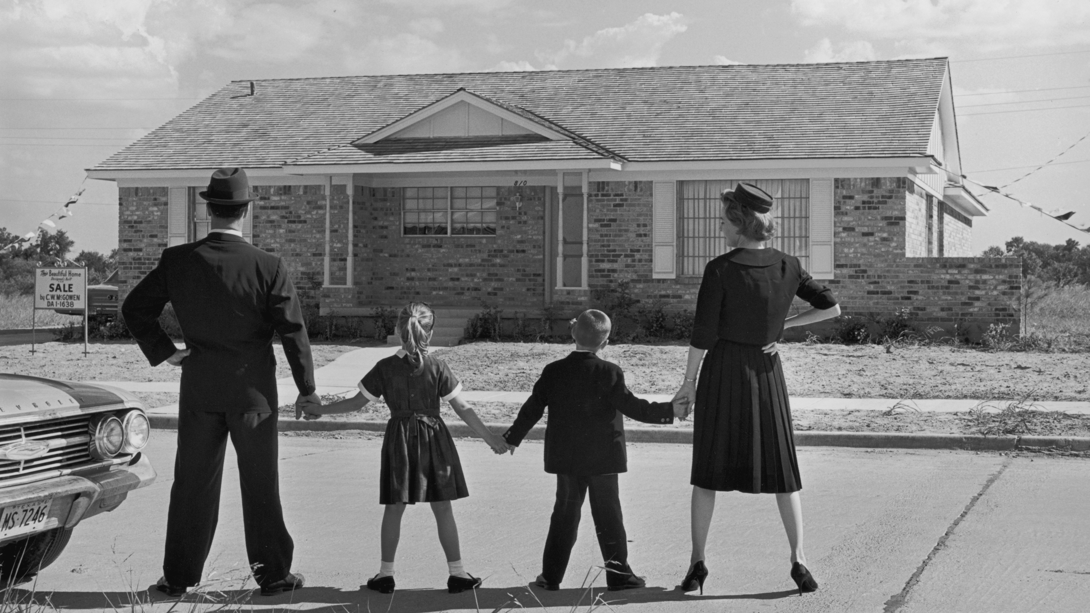 CIRCA 1965: Full-length image of a family of four holding hands while standing in a line with their backs to the camera, facing a ranch-style house in a suburban development. A sale sign is posted in the front yard of the house. (Photo by Camerique Archive/Getty Images)