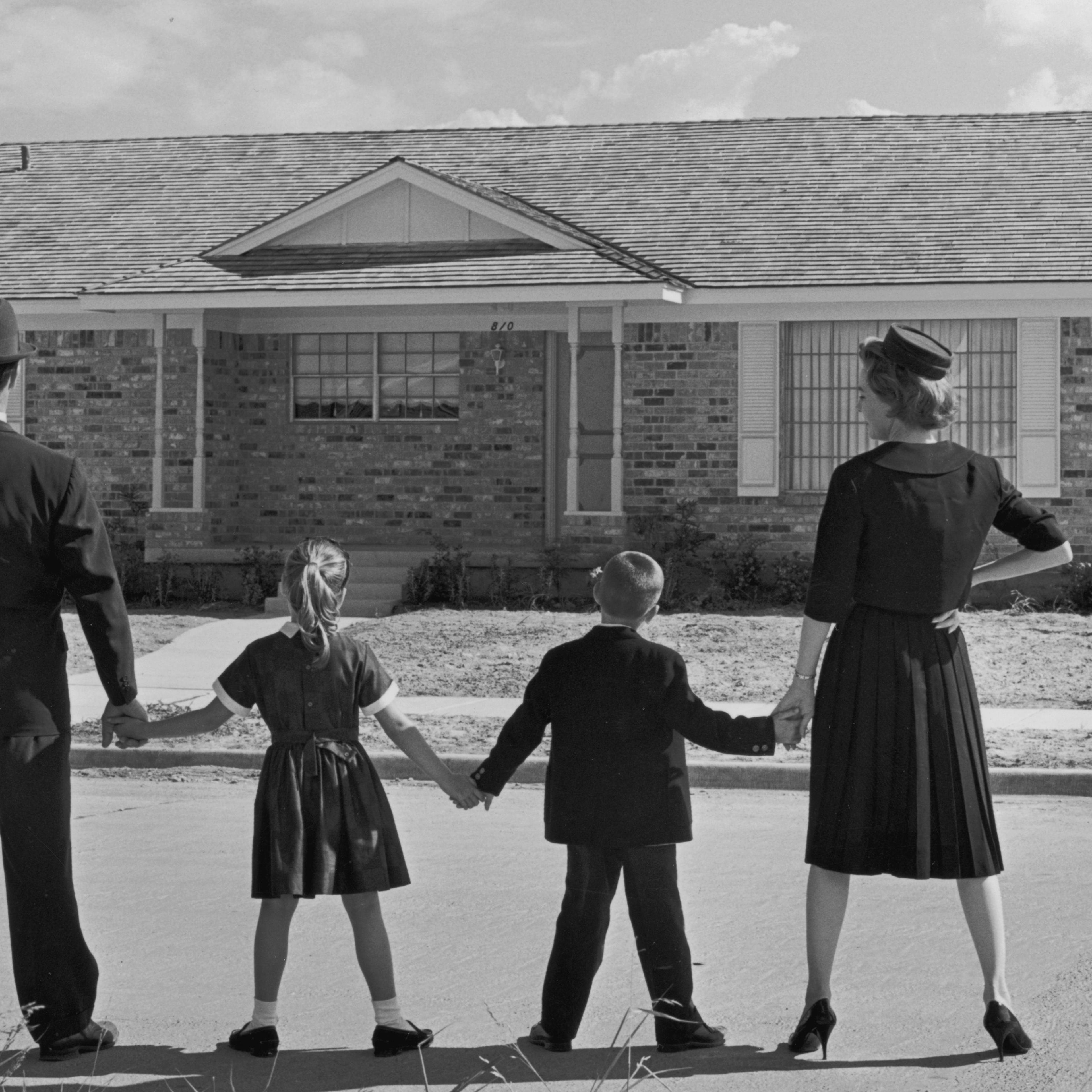 CIRCA 1965: Full-length image of a family of four holding hands while standing in a line with their backs to the camera, facing a ranch-style house in a suburban development. A sale sign is posted in the front yard of the house. (Photo by Camerique Archive/Getty Images)