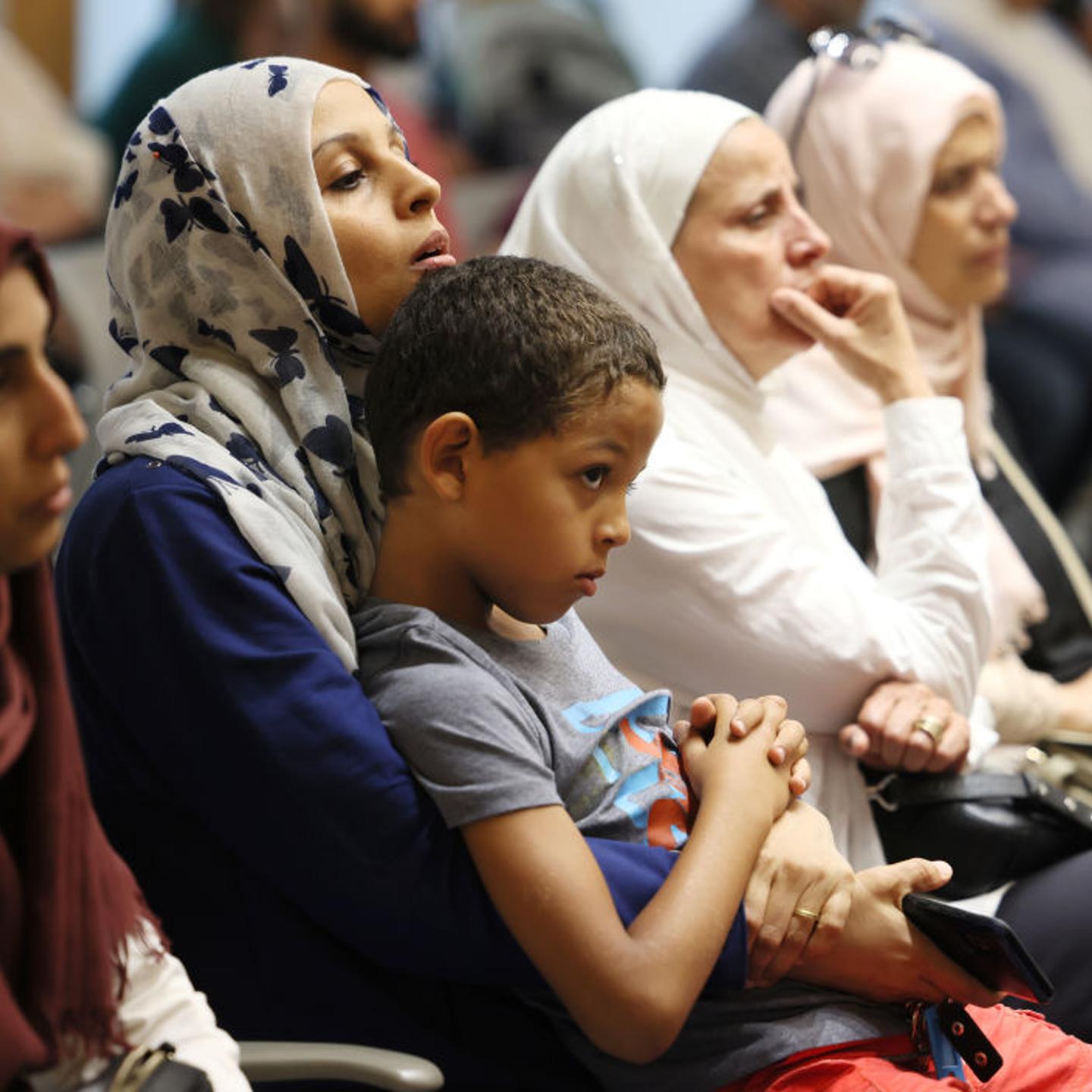 Muslim community members listen to a speaker during a prayer service and group meeting, with a mother holding a child on her lap.