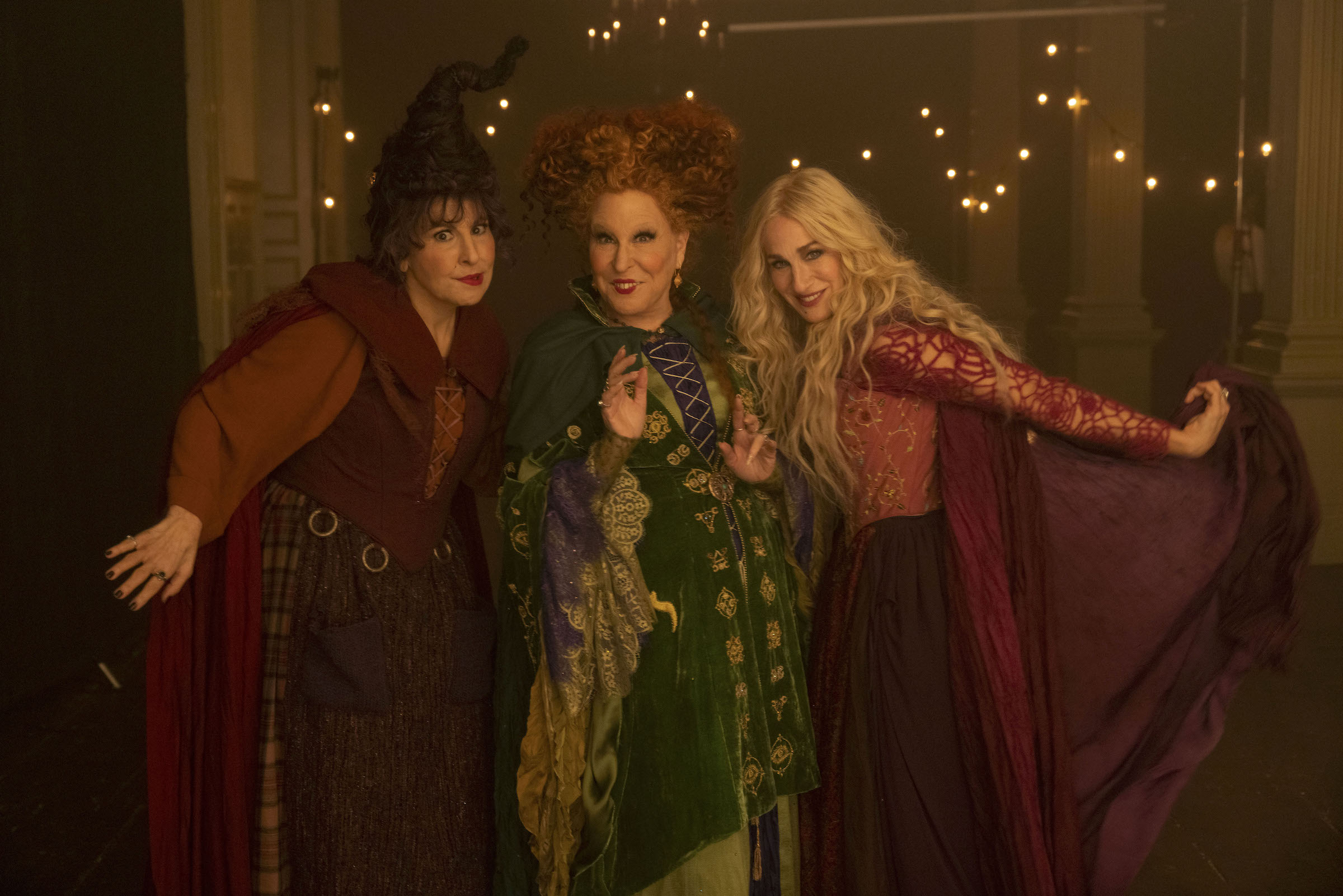 Three witches — two in burgundy/maroon capes and one in an olive green one — pose for the camera in front of fairy lights.