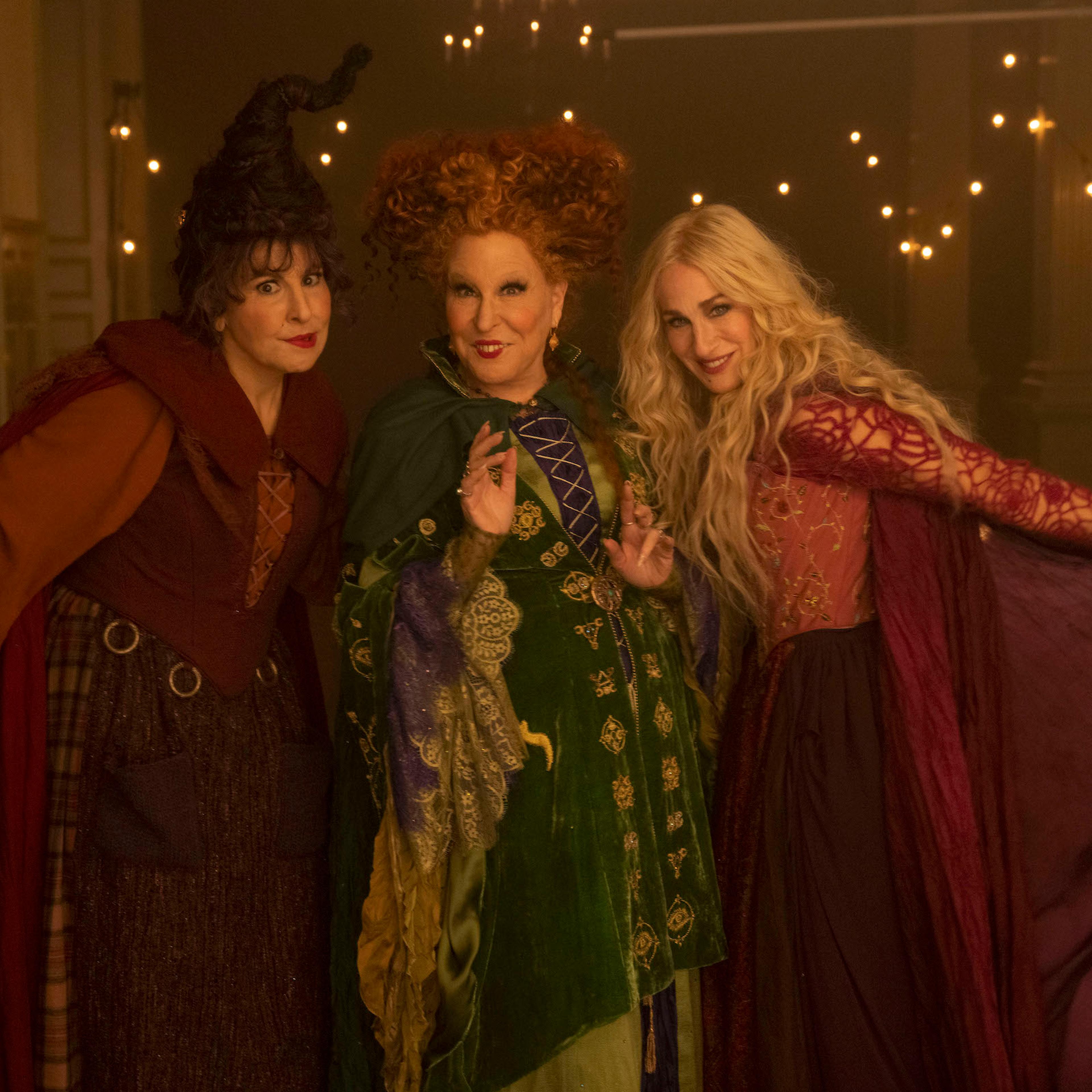 Three witches — two in burgundy/maroon capes and one in an olive green one — pose for the camera in front of fairy lights.