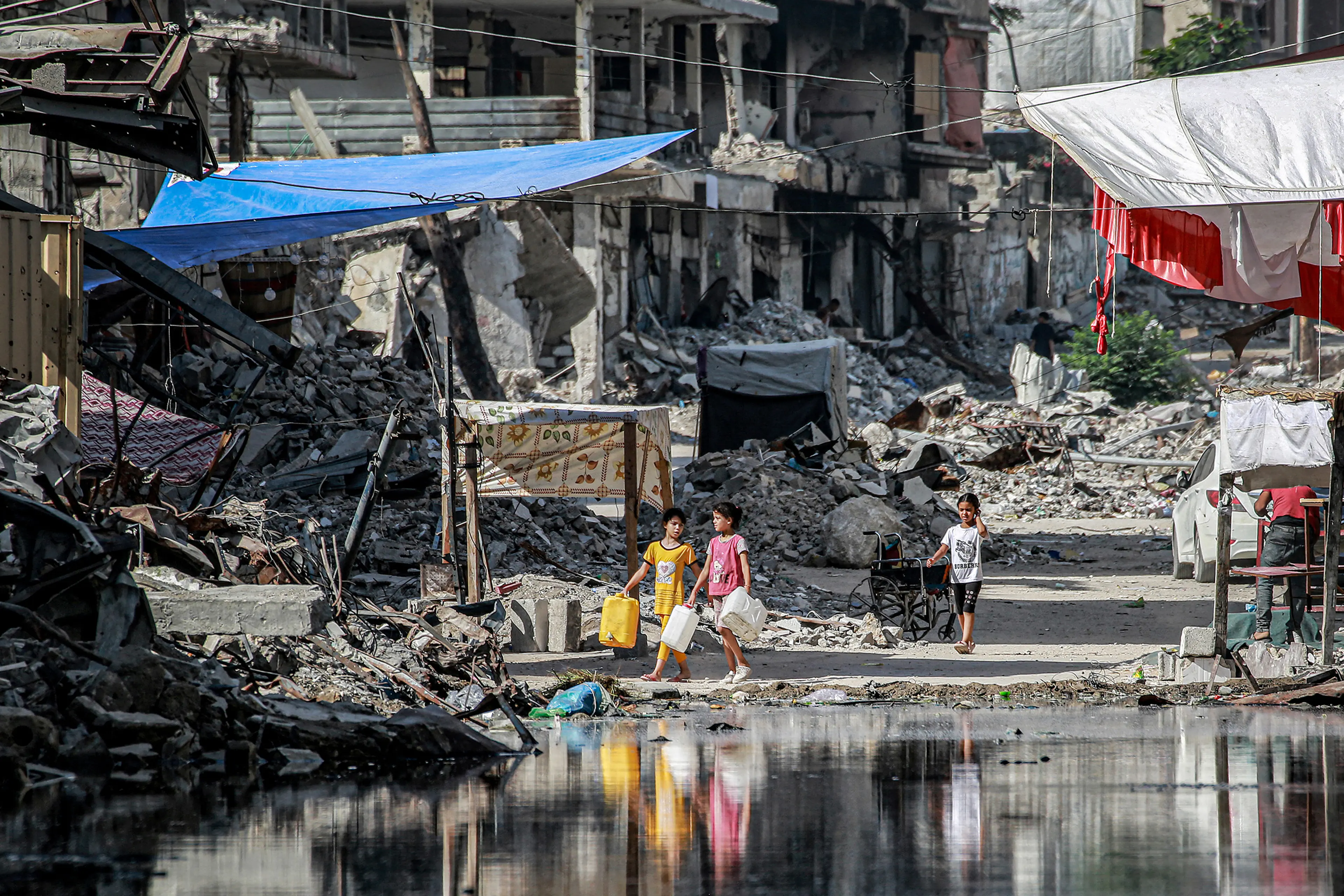 Girls walk with water containers past destroyed buildings and a puddle of sewage water erupting from collapsed underground pipes in Khan Yunis in the southern Gaza Strip on July 8, 2024.