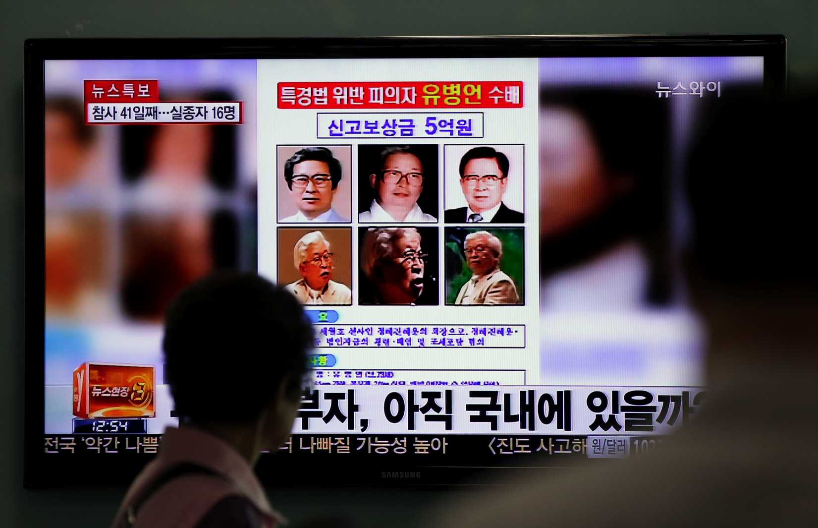 Commuters at a train station in Seoul walk past a screen displaying the wanted poster for Yoo Byung-eun and others, on Monday, May 26, 2014.