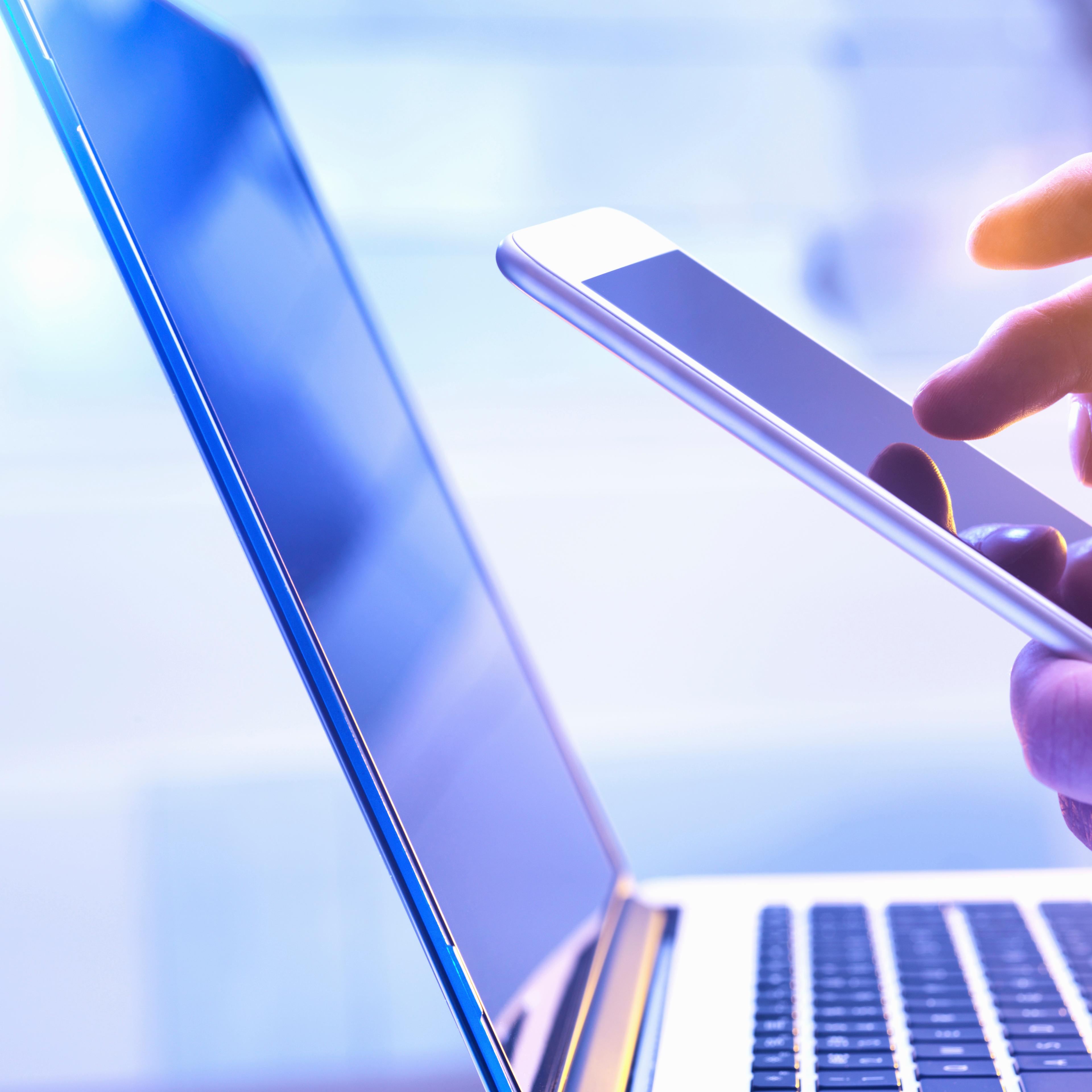 Man operating a touch screen mobile phone with a laptop in the background.