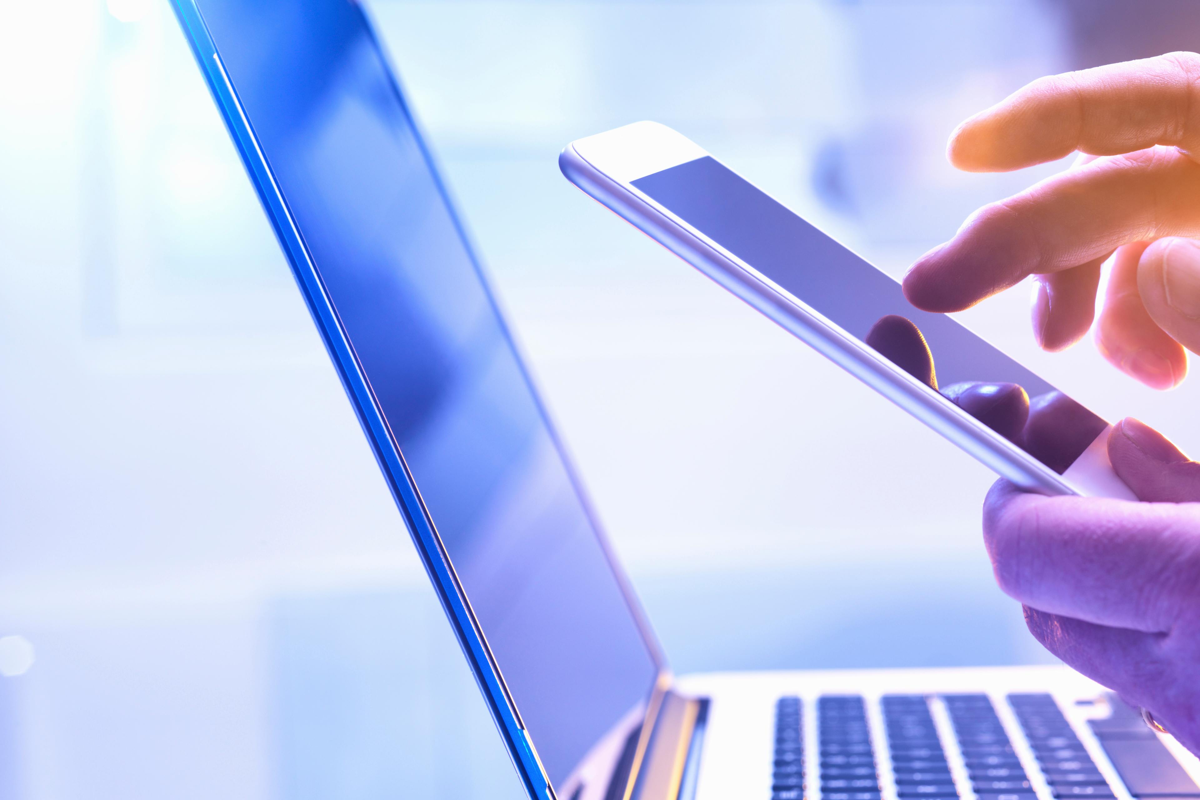 Man operating a touch screen mobile phone with a laptop in the background.