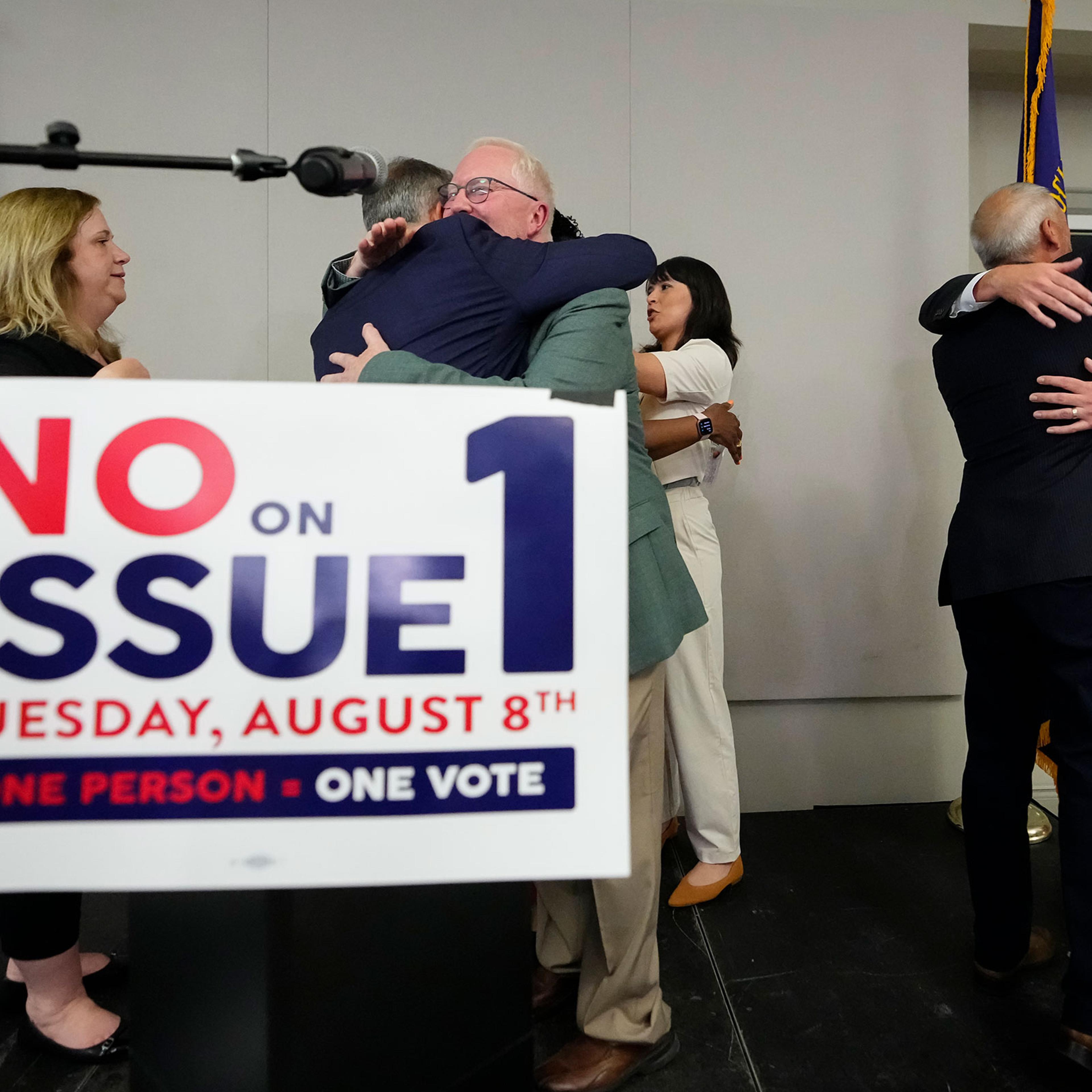 Organizers hug each other with a sign in front of them that says No on Issue 1