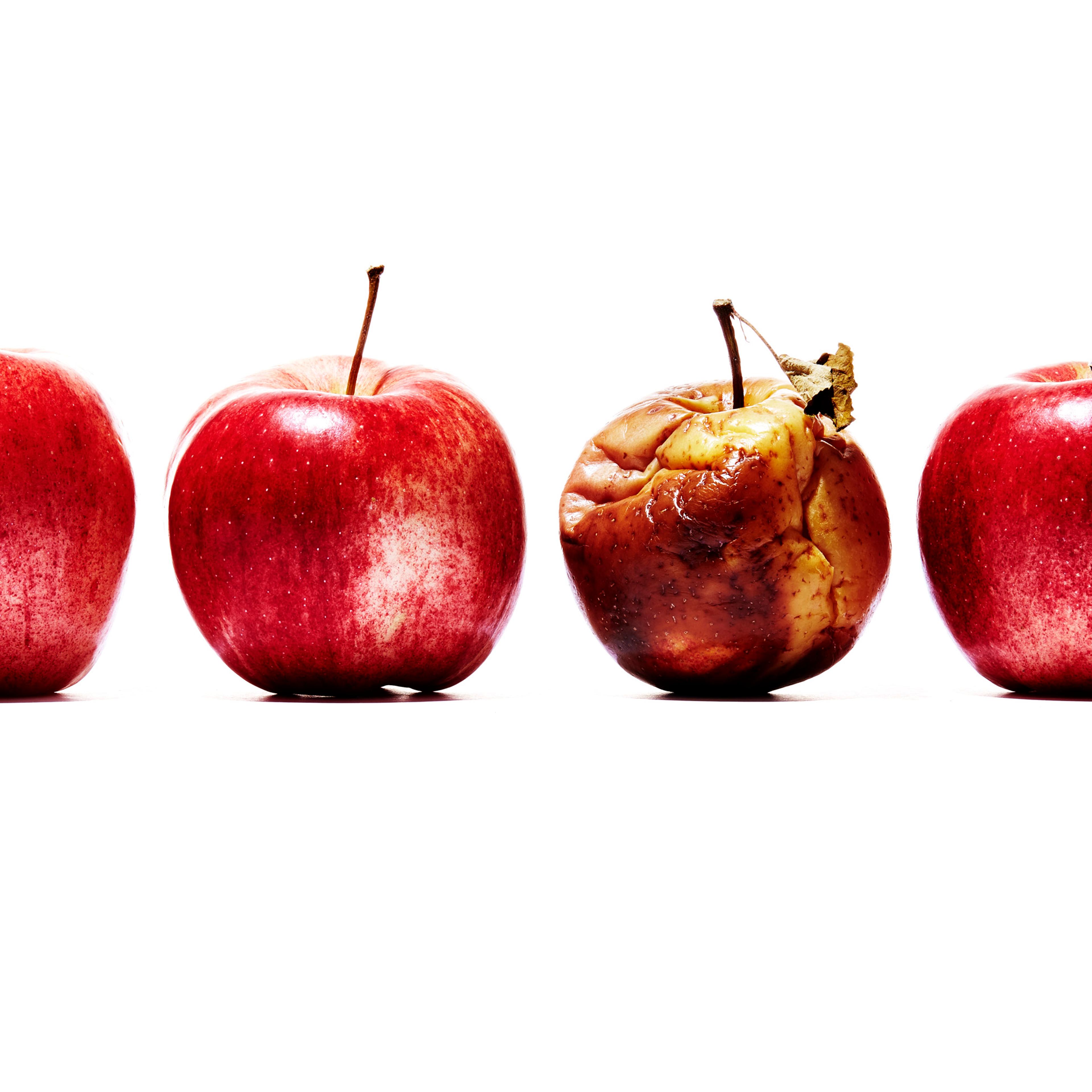 A still life showing 4 red apples in a row. The apple second from right is rotten.