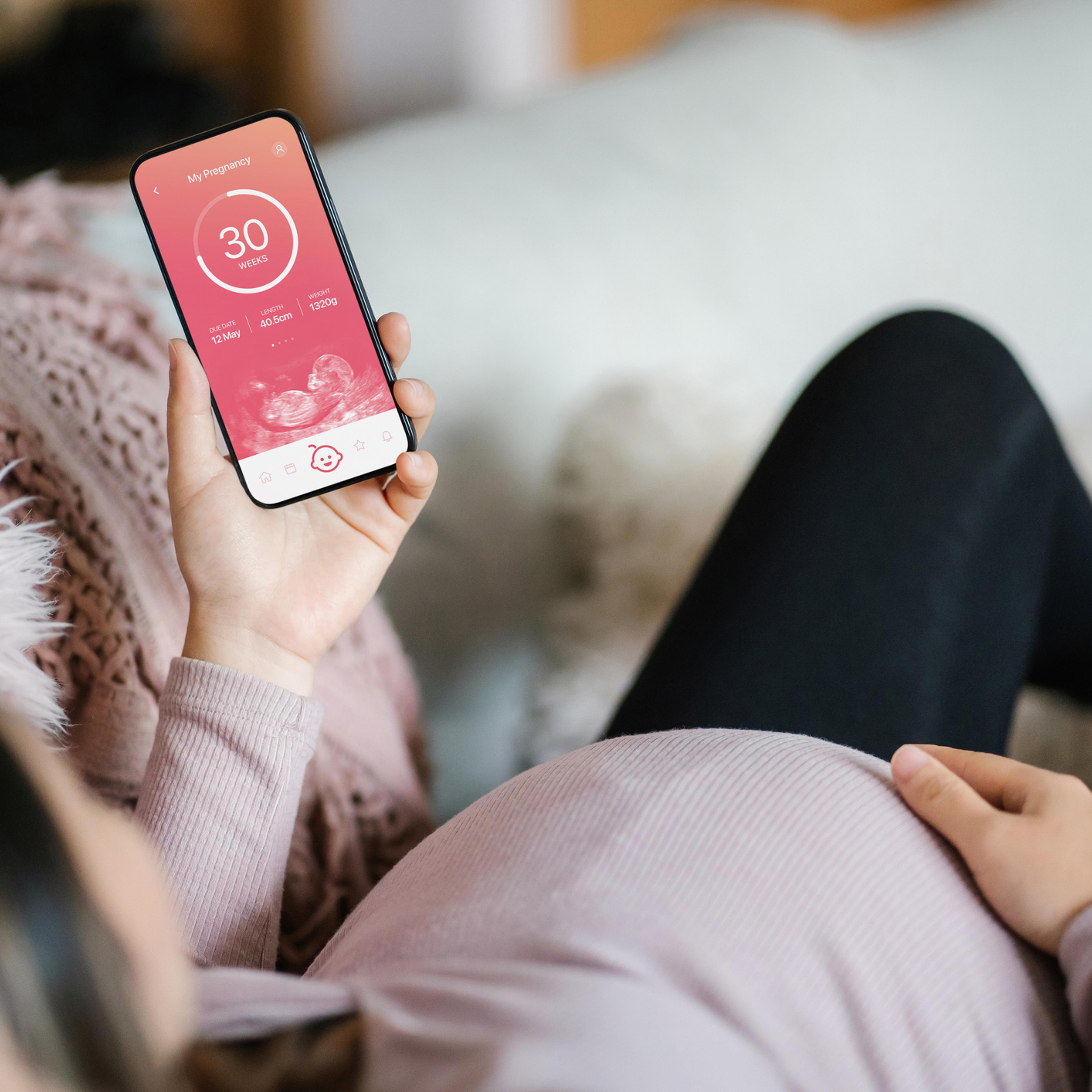Young Asian pregnant woman lying on sofa in the living room at cozy home, using baby tracker app on smartphone to monitor her baby's growth and development throughout the pregnancy journey. Technology and pregnancy lifestyle