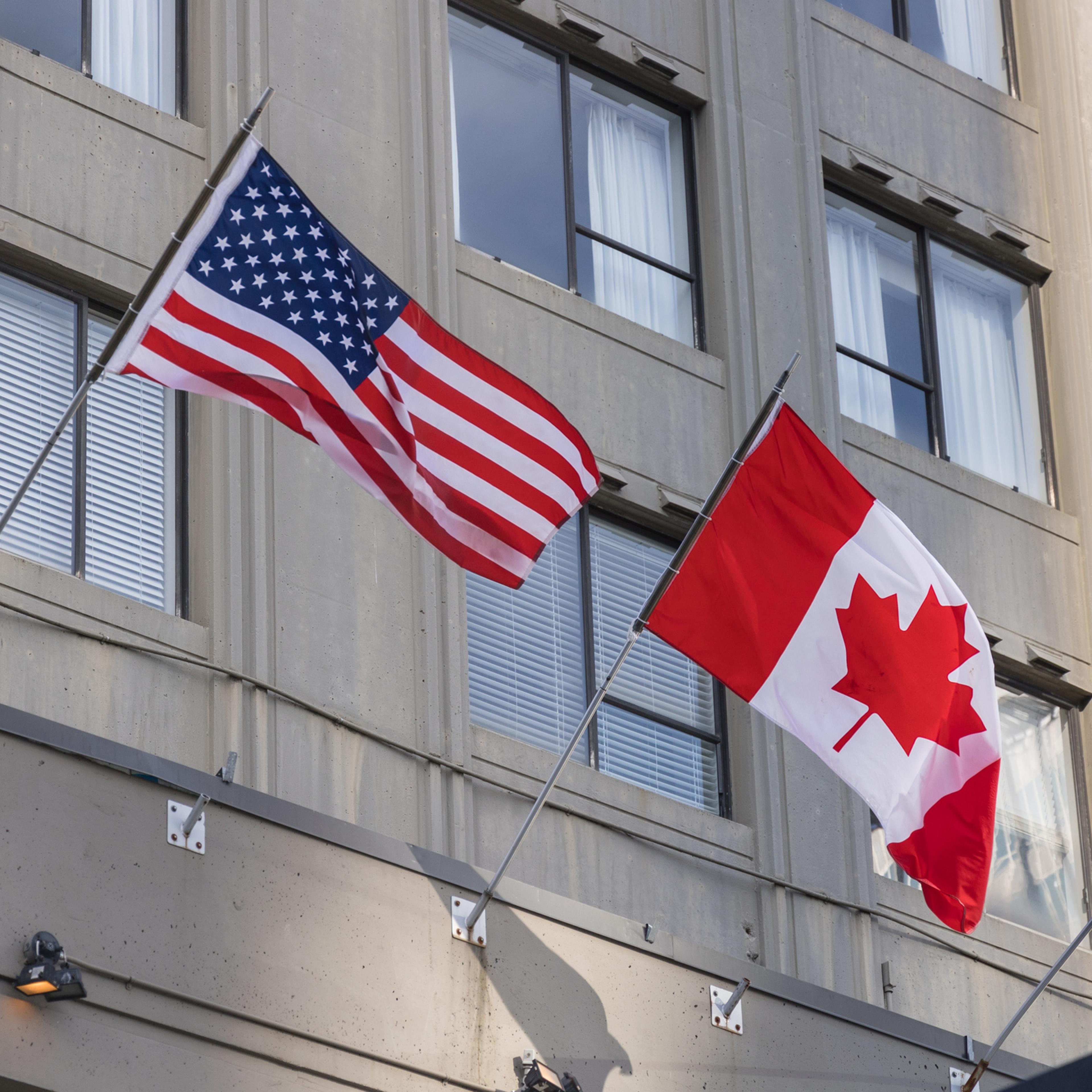 Flags of the United States and Canada waving against a building facade.