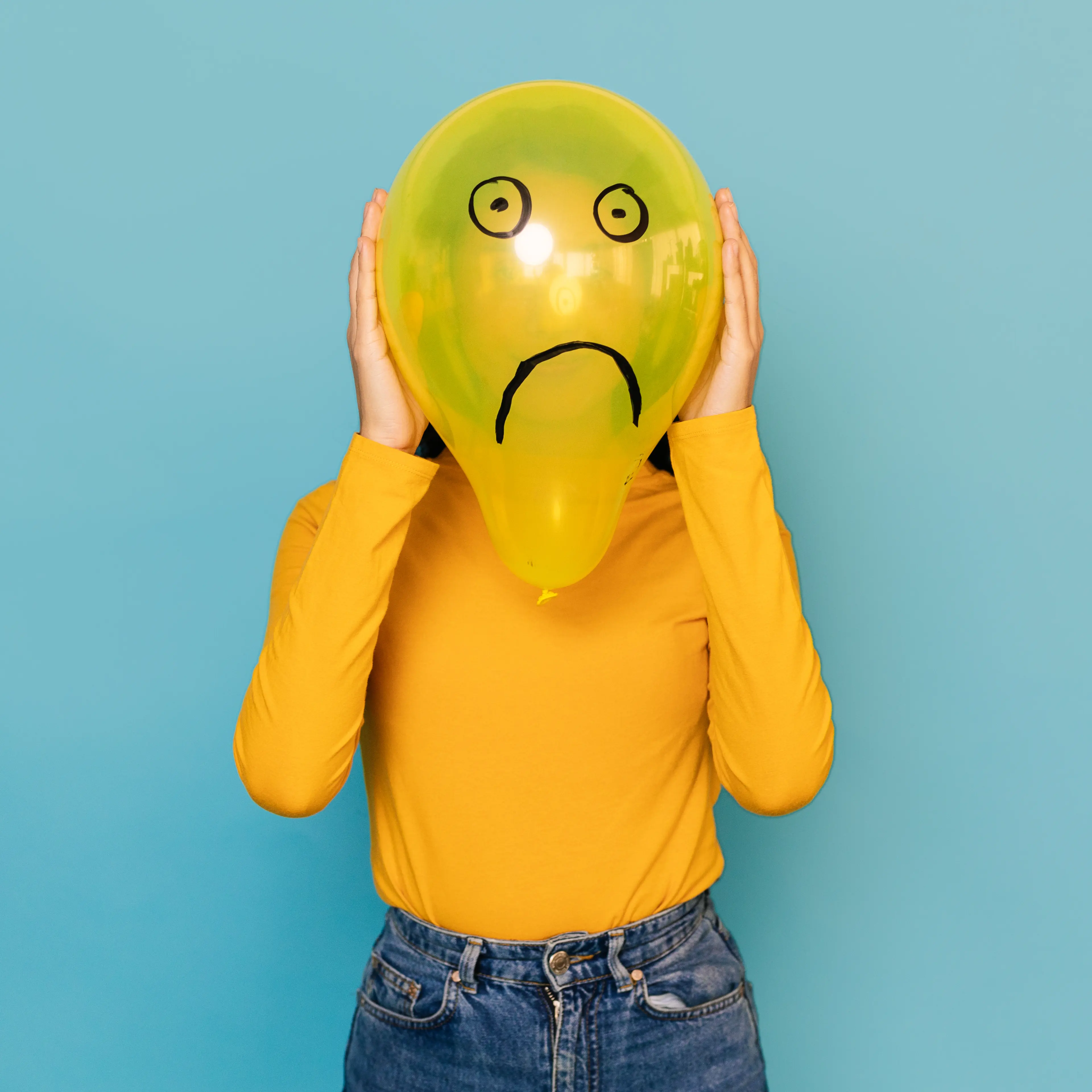 Young woman hidden behing a balloon with a sad face drawn on it over blue background. Negative emotion concept