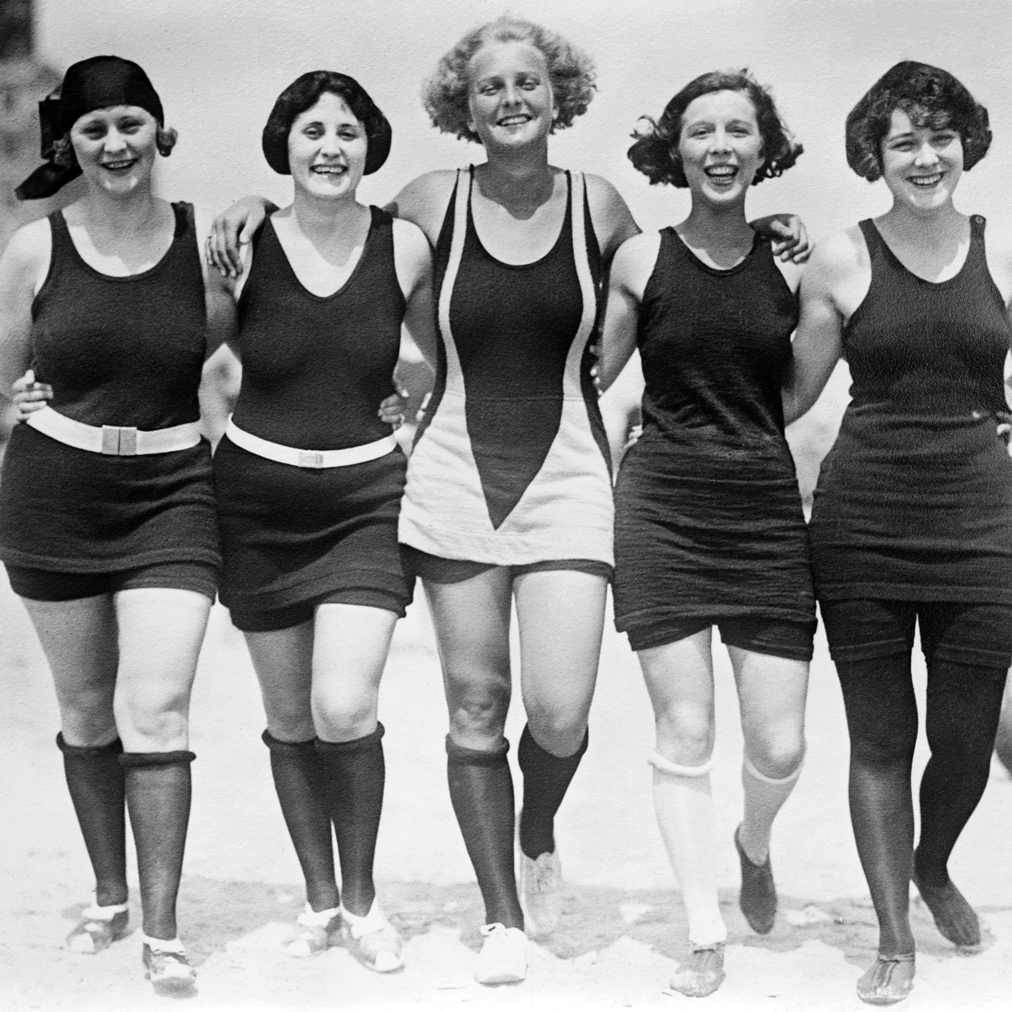 Five women walking arm-in-arm on the beach wearing wool bathing suits, circa 1925.