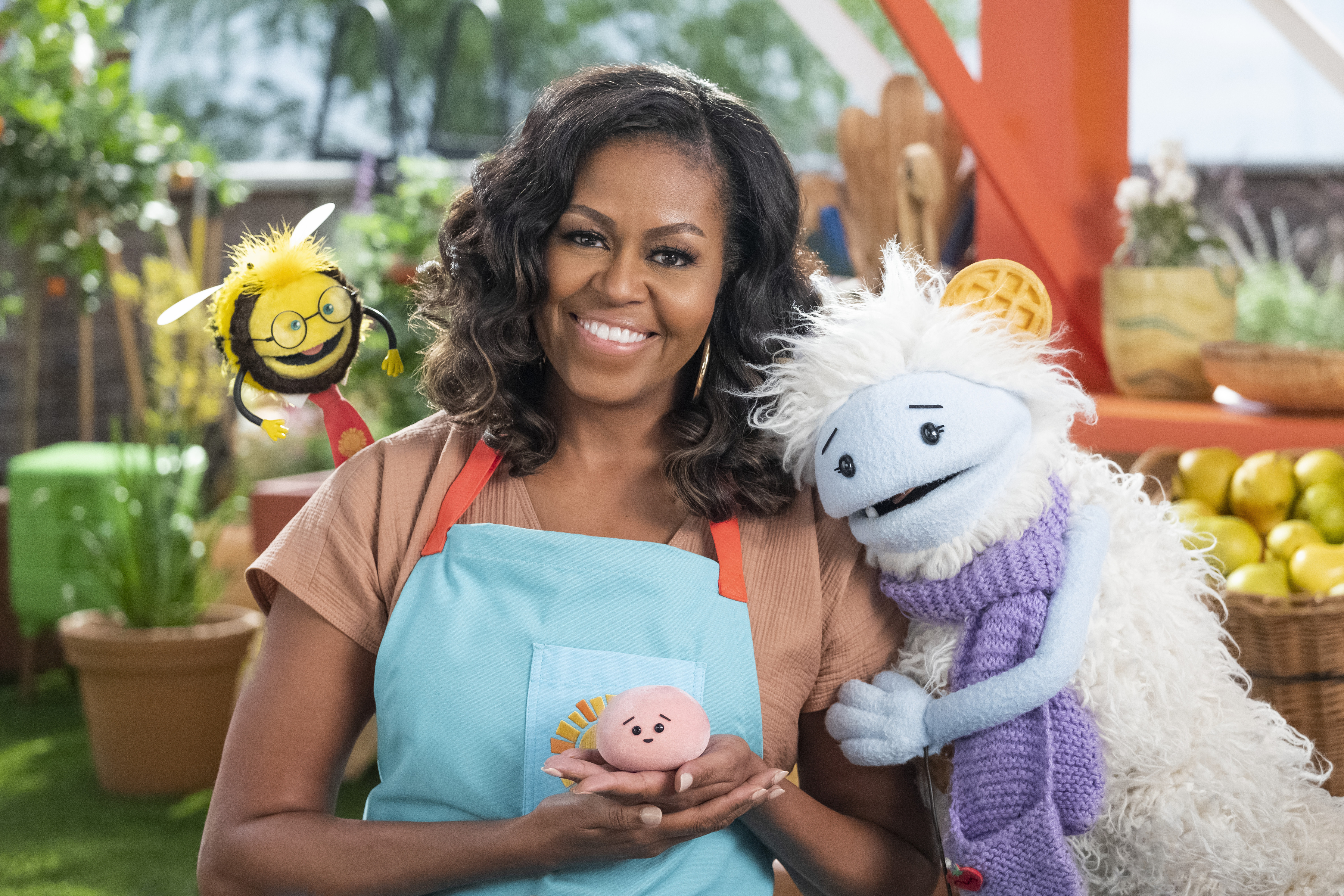 Michelle Obama, wearing a blue apron, stands in a rooftop garden posing for the camera and holding a pink, round mochi puppet. A bee puppet wearing a red tie hovers over her shoulder while a furry white and blue puppet with frozen waffle ears embraces he