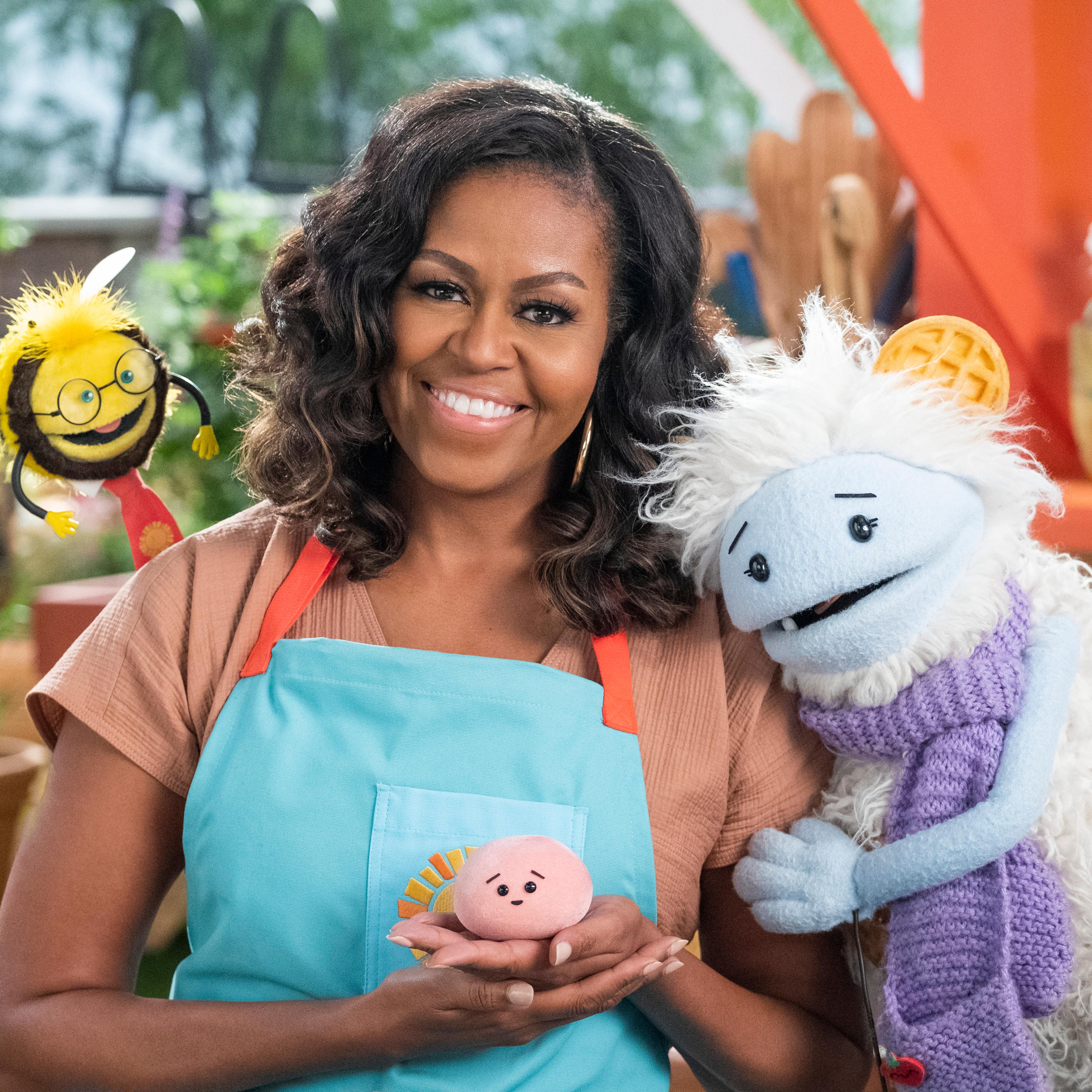 Michelle Obama, wearing a blue apron, stands in a rooftop garden posing for the camera and holding a pink, round mochi puppet. A bee puppet wearing a red tie hovers over her shoulder while a furry white and blue puppet with frozen waffle ears embraces he