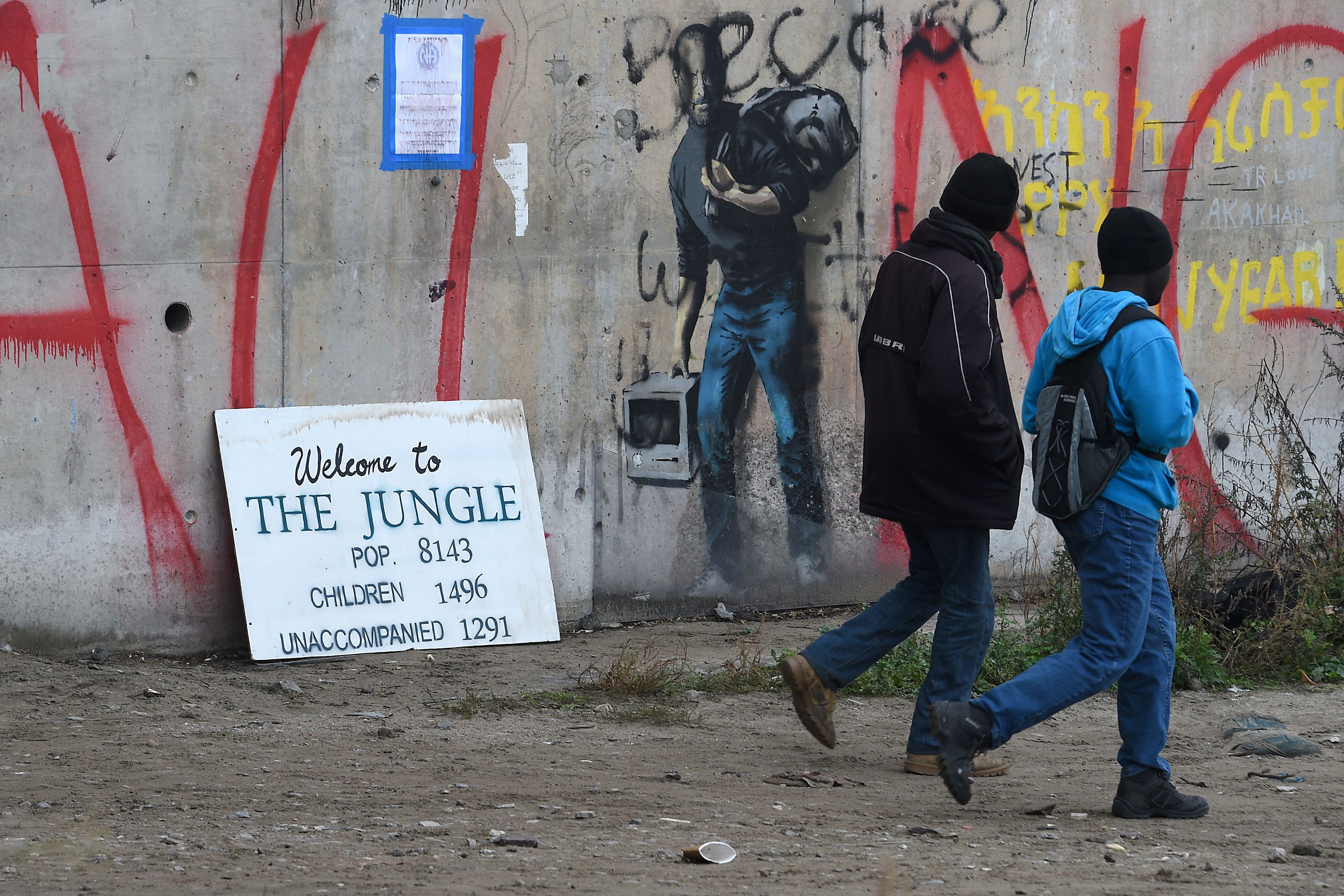 Migrants walk past a graffiti by street artist Banksy representing Apple founder Steve Jobs as a migrant during the full evacuation of the Calais "Jungle" camp, in Calais, northern France, on October 24, 2016.