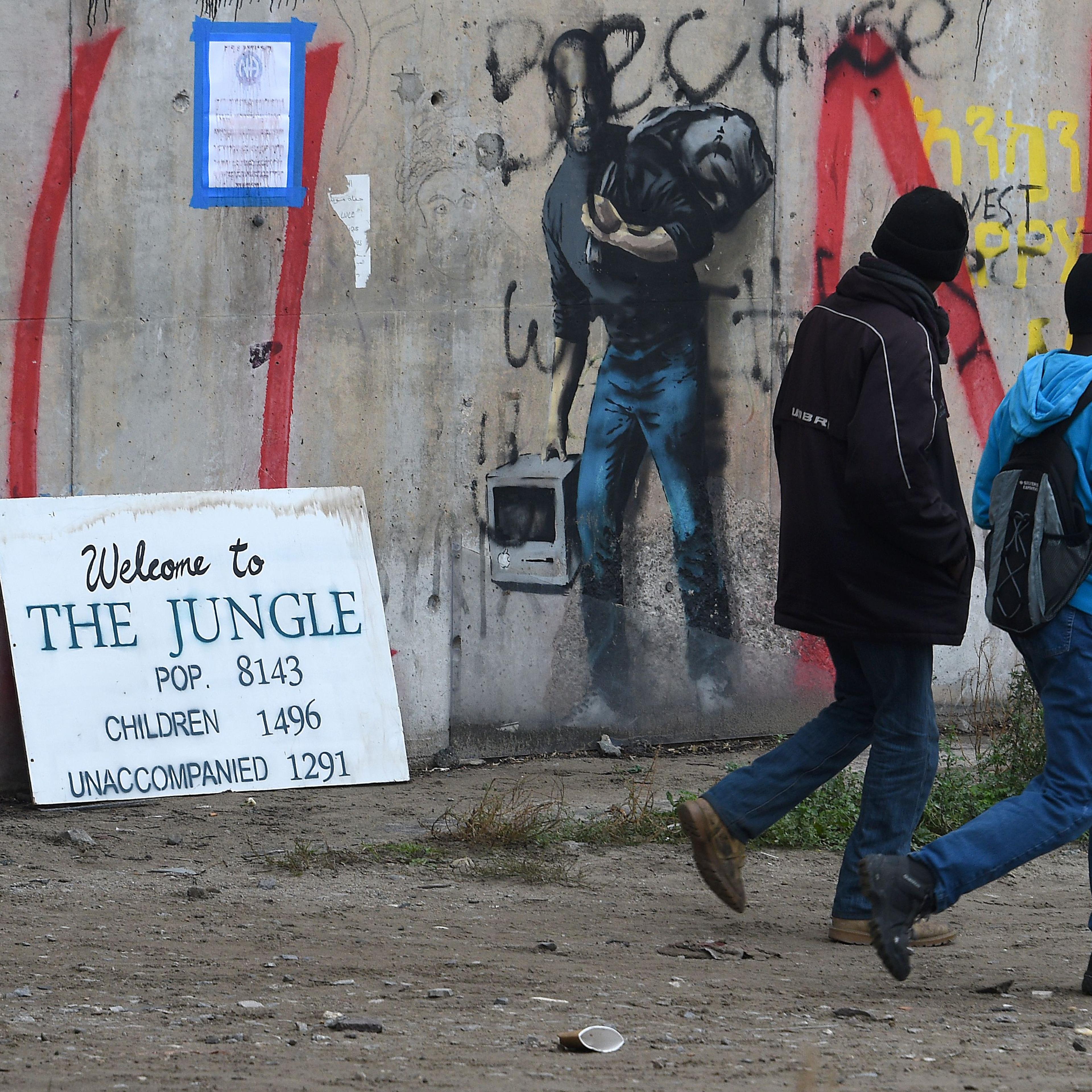 Migrants walk past a graffiti by street artist Banksy representing Apple founder Steve Jobs as a migrant during the full evacuation of the Calais "Jungle" camp, in Calais, northern France, on October 24, 2016.