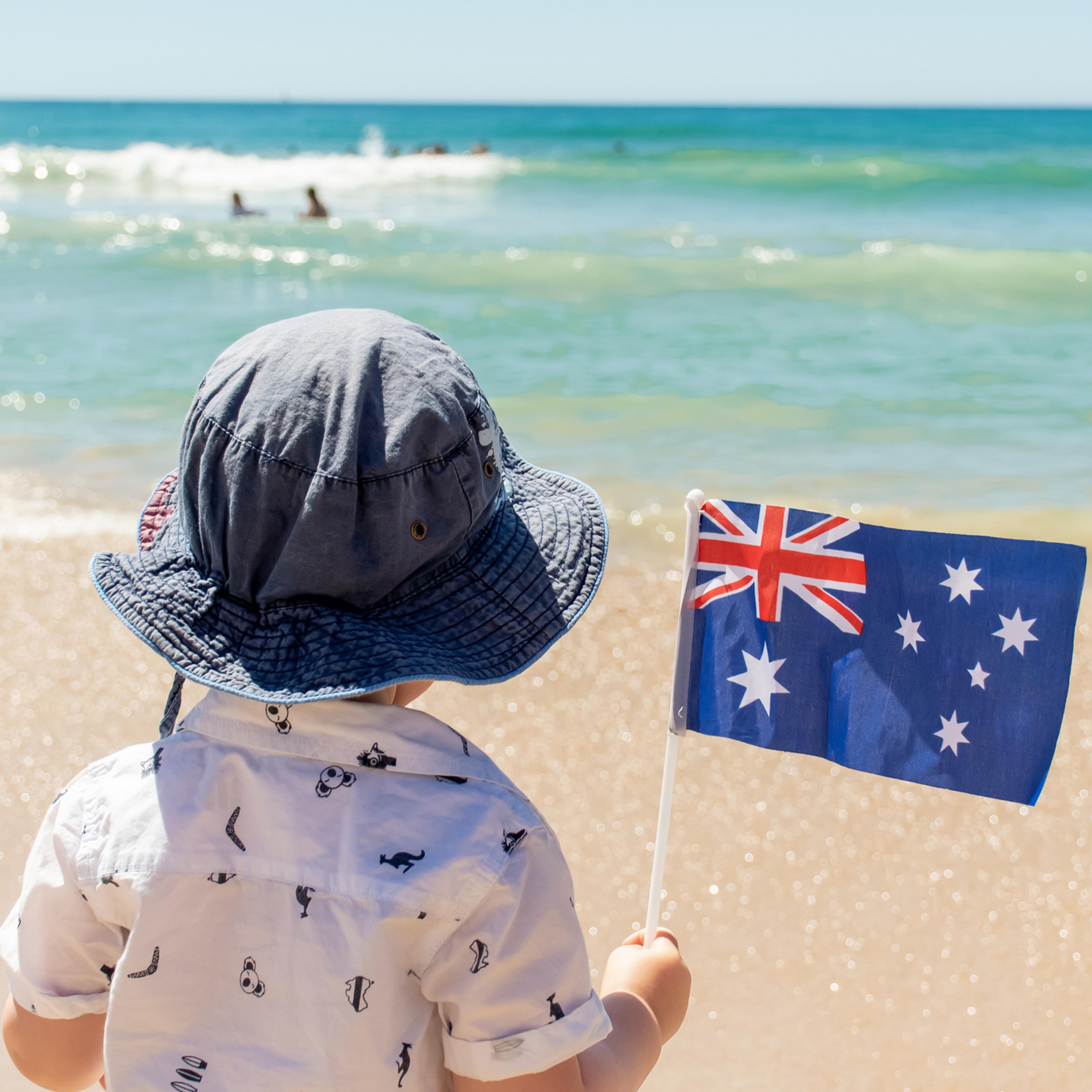 Little boy wearing hat holding Australian flag on a sandy ocean beach. Australia Day concept