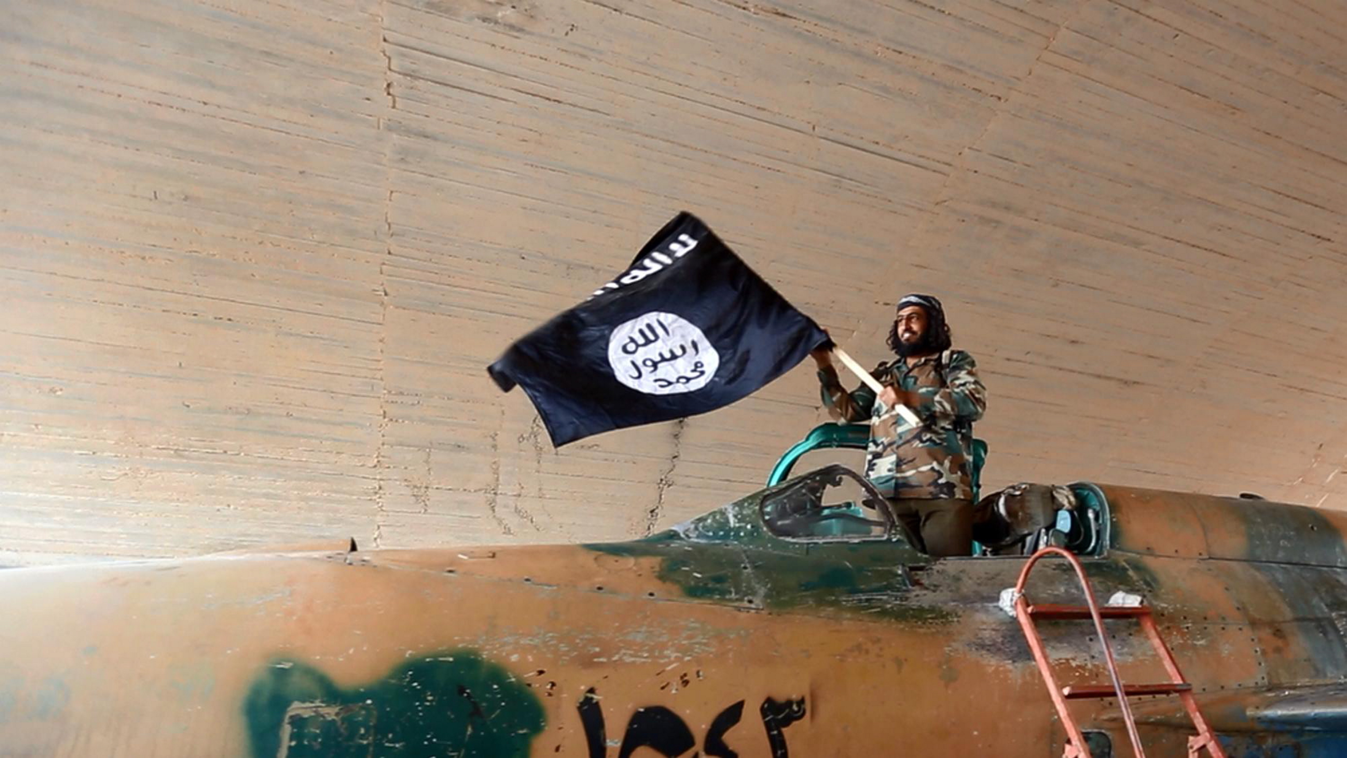 ISIS fighter waves a flag while standing on a captured government fighter jet in Raqqa, Syria, 2015.