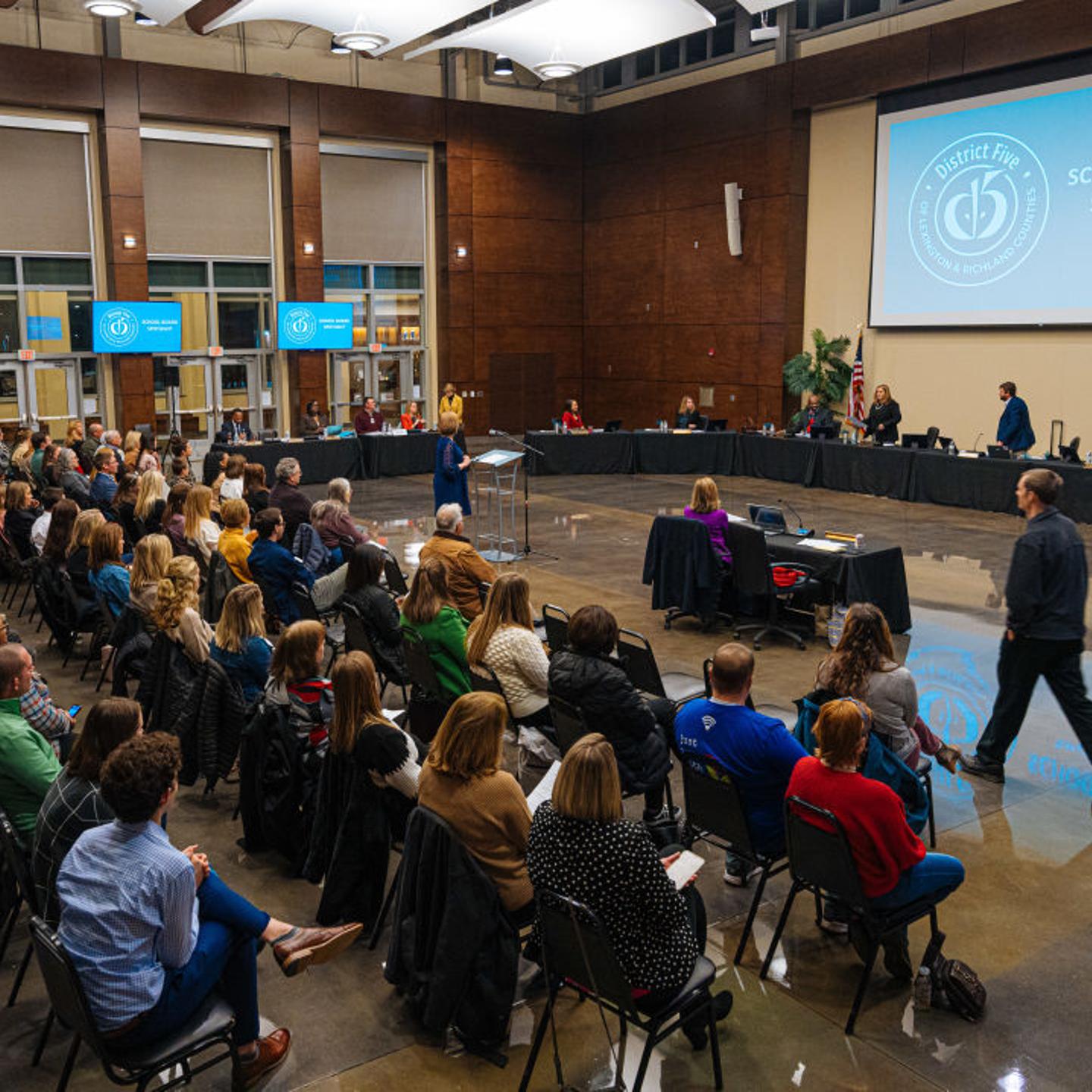 A school board is called into session in front of several people seated in chairs.