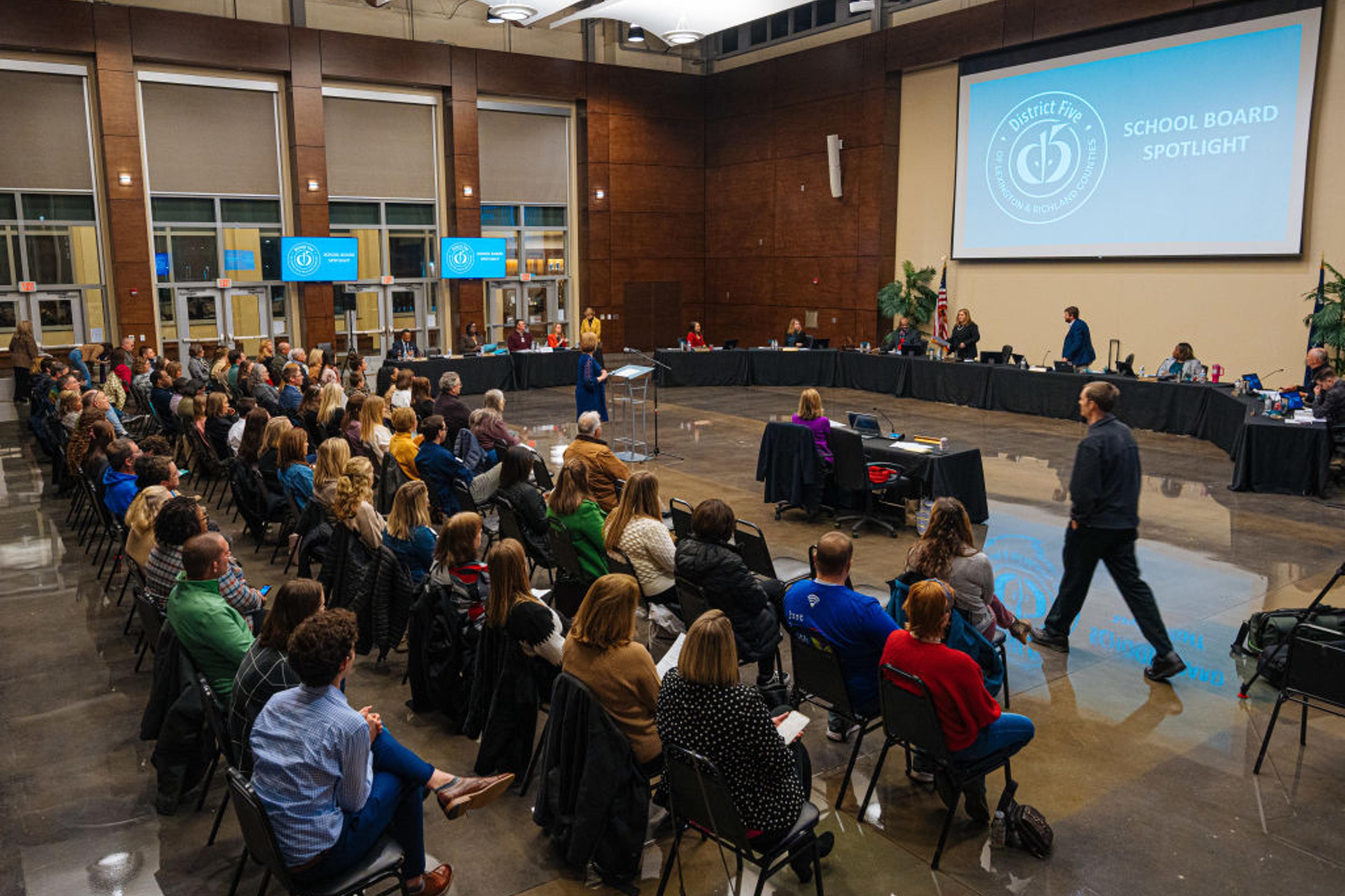 A school board is called into session in front of several people seated in chairs.
