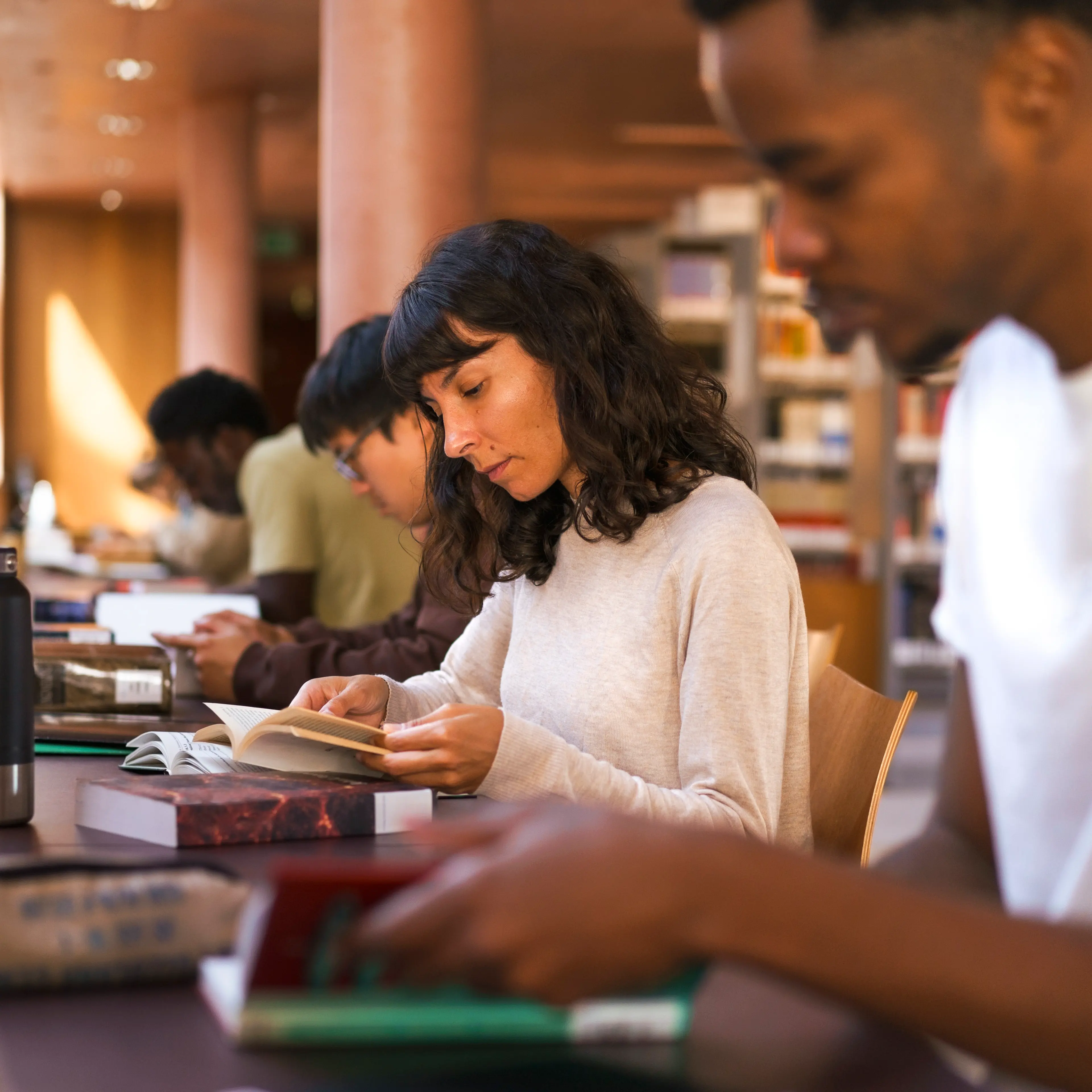 Confident female student reading book while sitting at desk in college library
