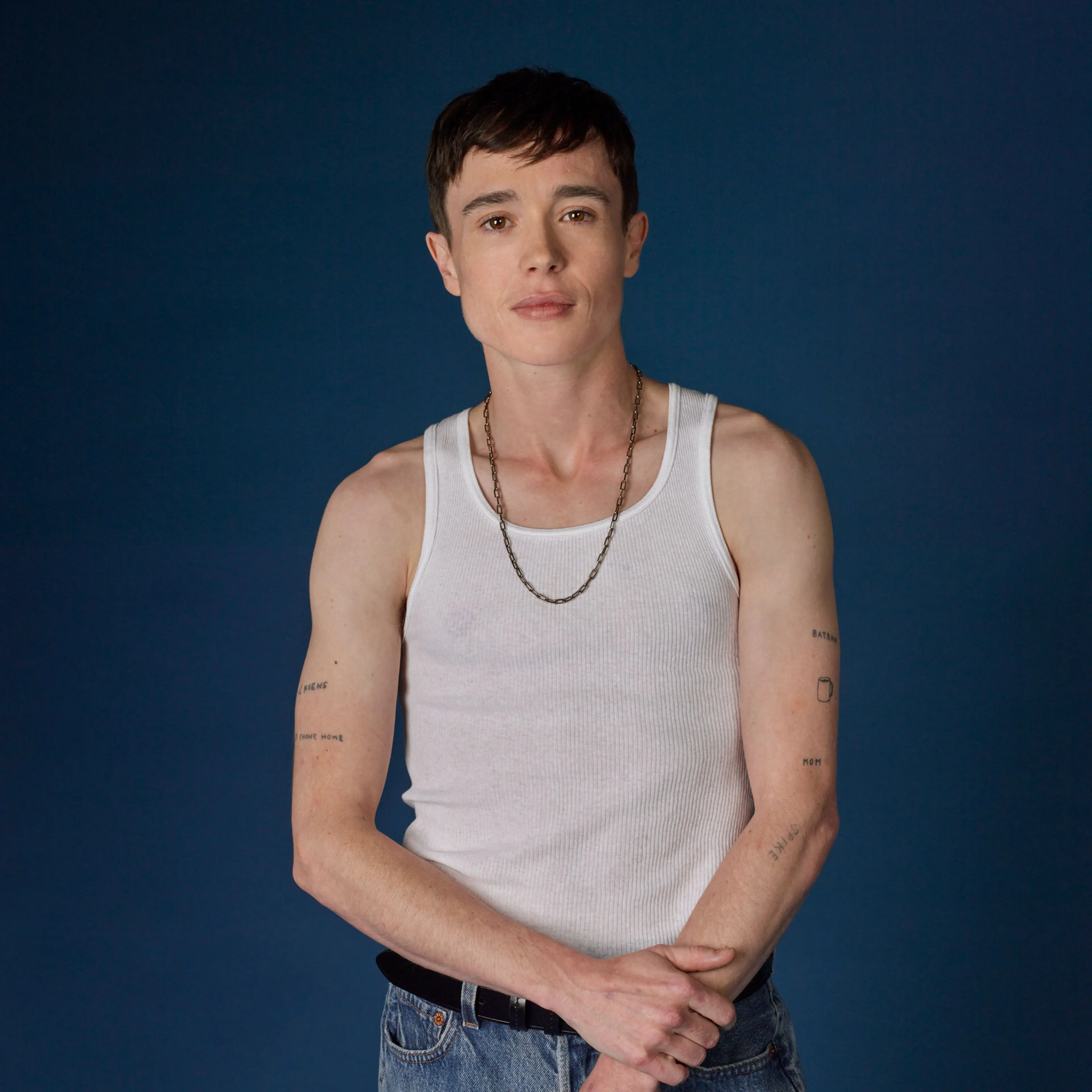 Elliot Page wearing a white tank and jeans, posing against blue background