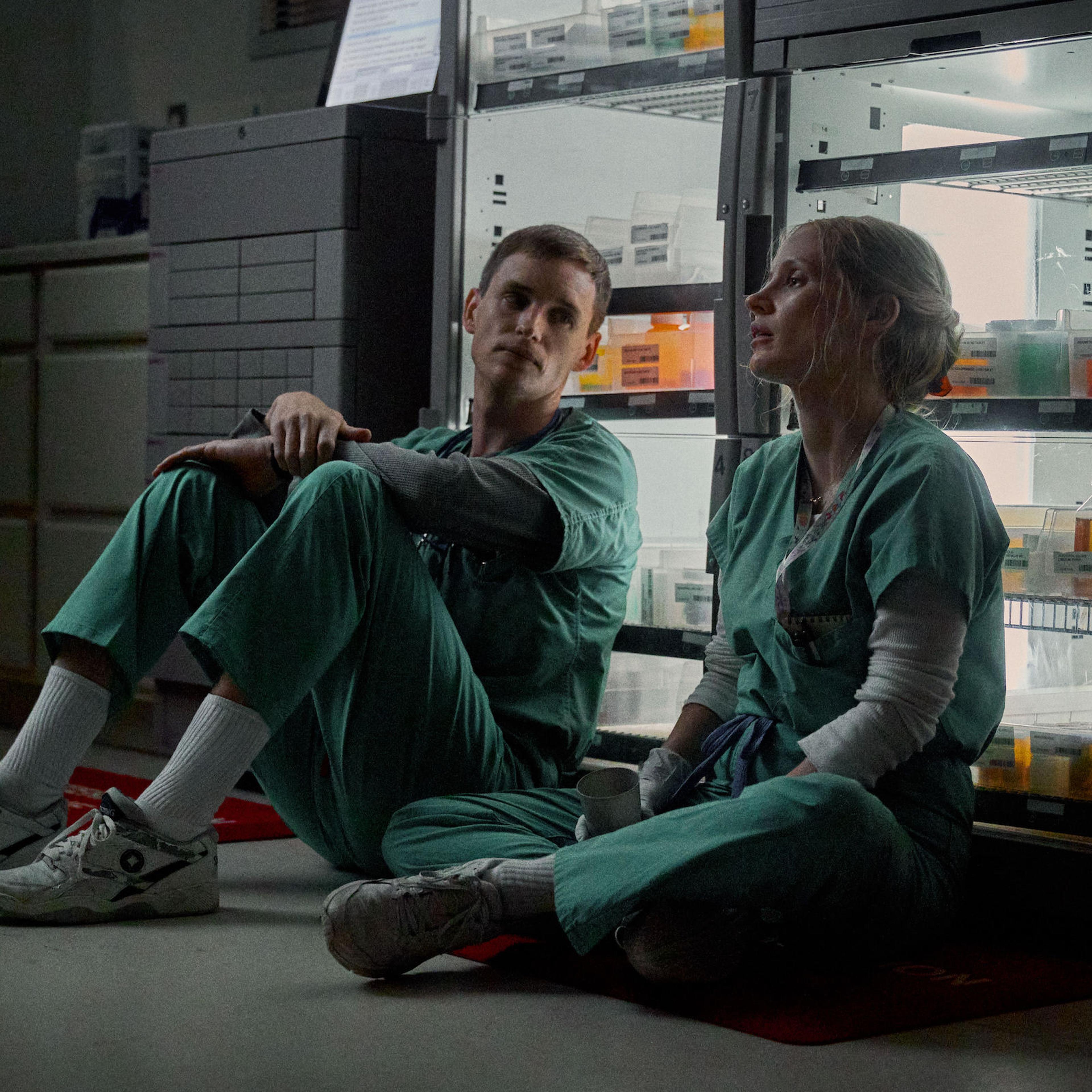 Charlie and Amy sit side-by-side on the floor, leaning up against a fridge full of medical samples