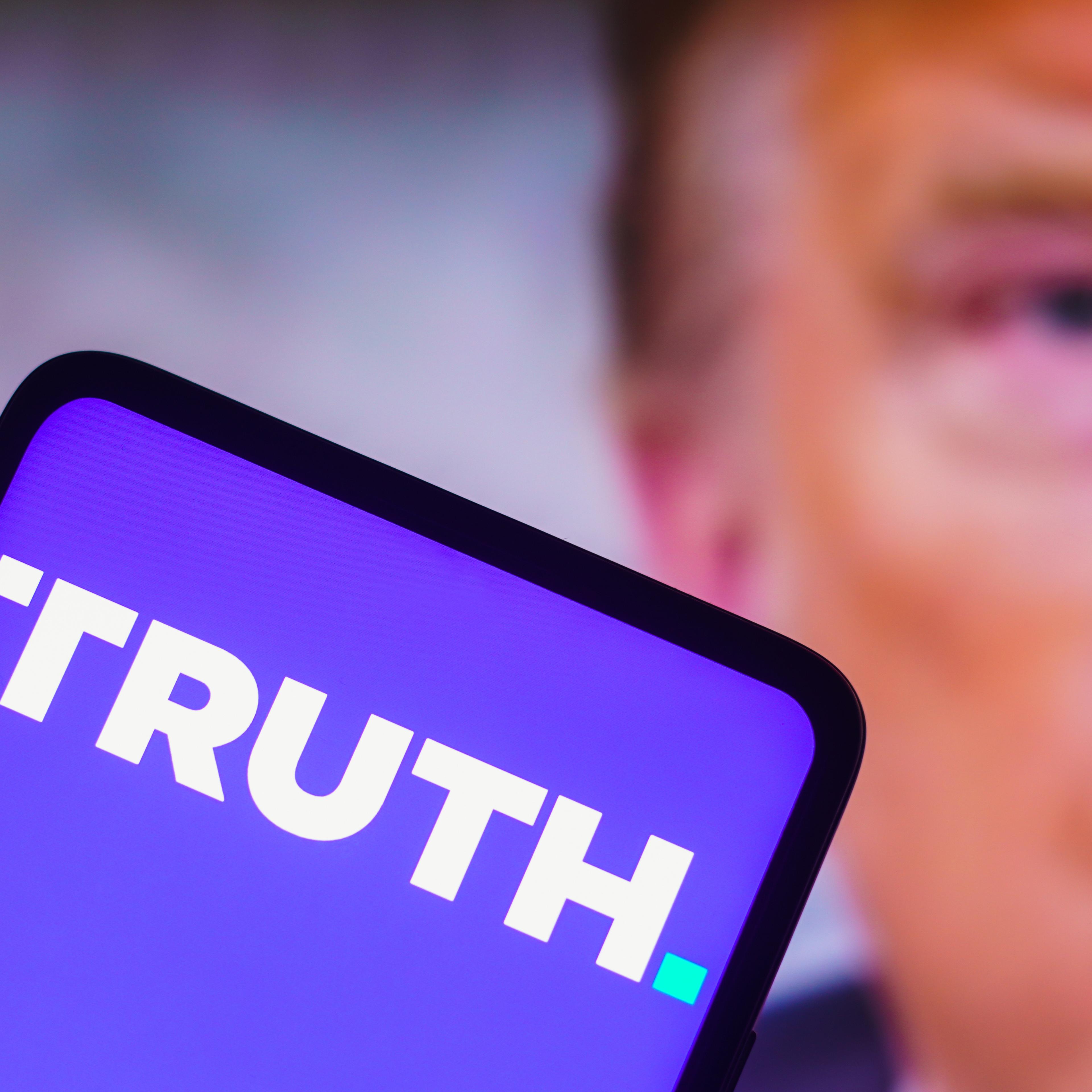 In this photo illustration, the TRUTH Social logo is seen displayed on a smartphone with a photo of former U.S. President Donald Trump in the background.
