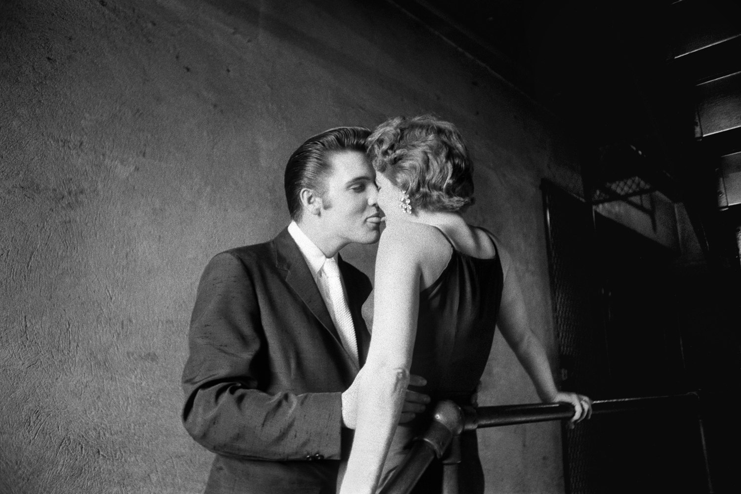 June 30, 1956. Elvis with Bobbi Owens, backstage at the Mosque Theatre, Richmond, Virginia. Elvis had one  of his bodyguards drive from Memphis to South Carolina [400 mikes], pick up Bobbi and bring her up to Richmond. Elvis meets her at the hotel, then he takes her to the theater. After Elvis finishes combing his hair, he disappears on me. I walk down the stairwell … and I see two figures at the end of the hallway ... and I’m thinking about what Capa said, that if you aren’t close enough your photos are probably boring. So I’m going down on the landing and no sooner do I get myself set when she says to him, 'Elvis, I bet you can’t kiss me?' That’s all he needed, so he said, 'I betcha I can.' Two weeks later I developed my film in my laboratory [and realize he'd tried to kiss her twice]. He bent her nose the first time, but the second time it was perfect, tongue to tongue, tip to tip, and [55 years later] she denies on national television that he ever did kiss her… that she was really