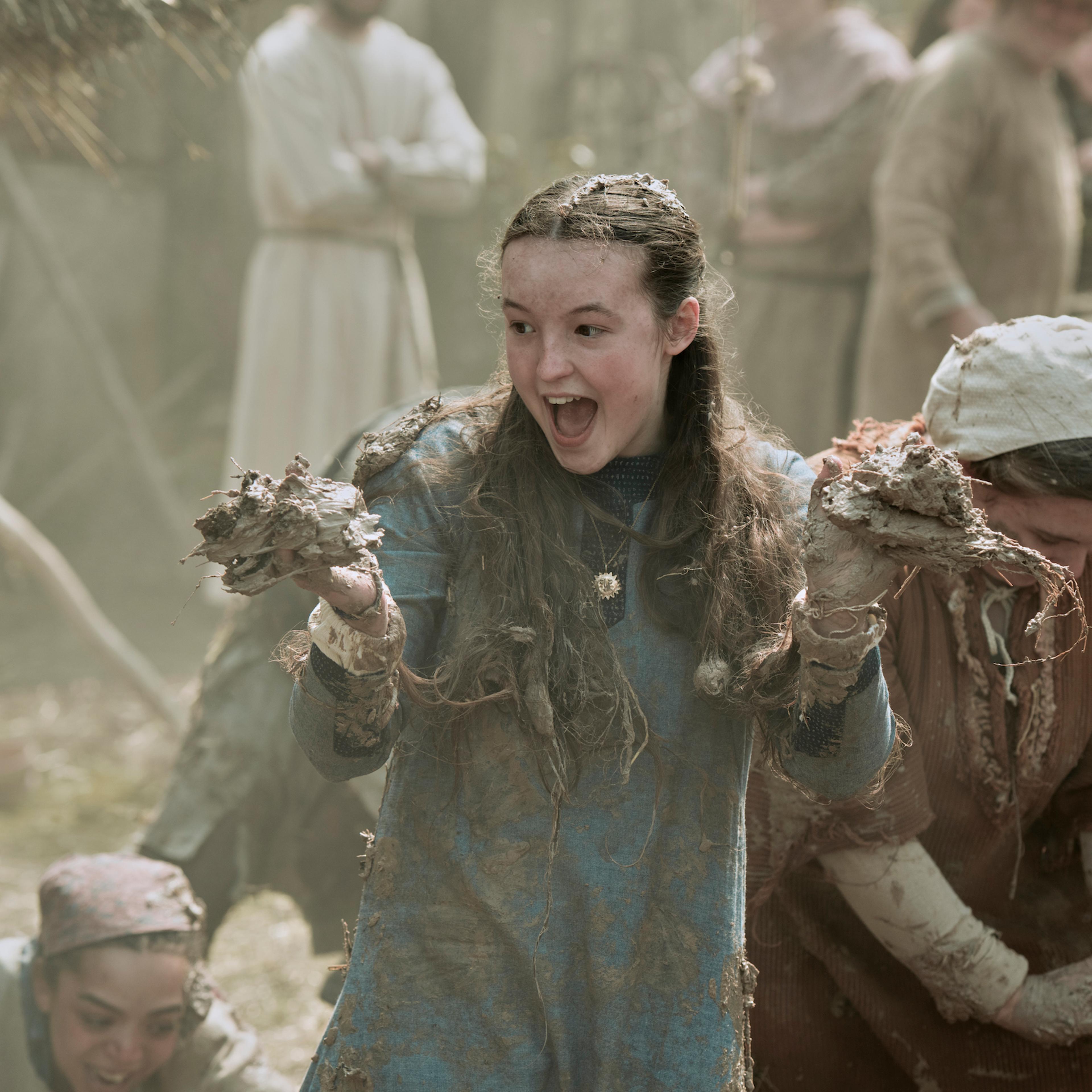 Birdy, in a longsleeve blue dress, grabs fistfuls of mud at a village cottage raising
